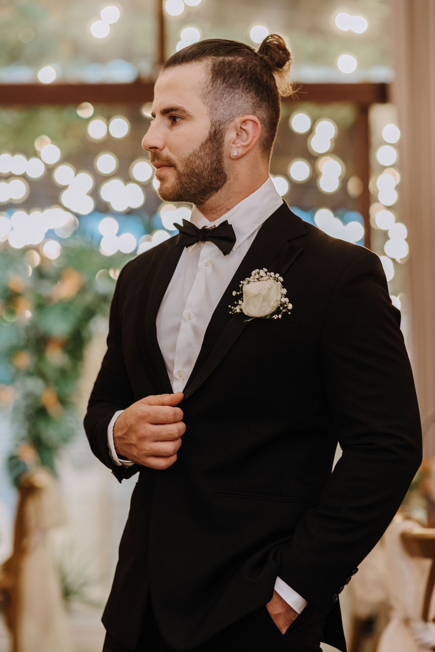 A groom in a black tuxedo with a bow tie and boutonniere stands in front of twinkling lights.