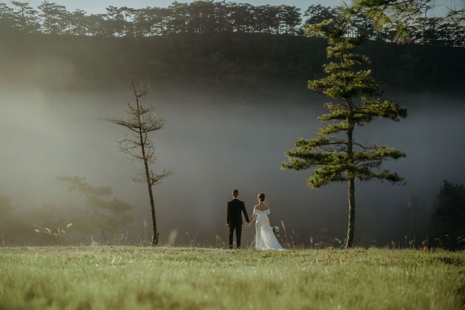 Two people stand together on a misty hillside with sparse pine trees in Da Lat, Vietnam during sunset.