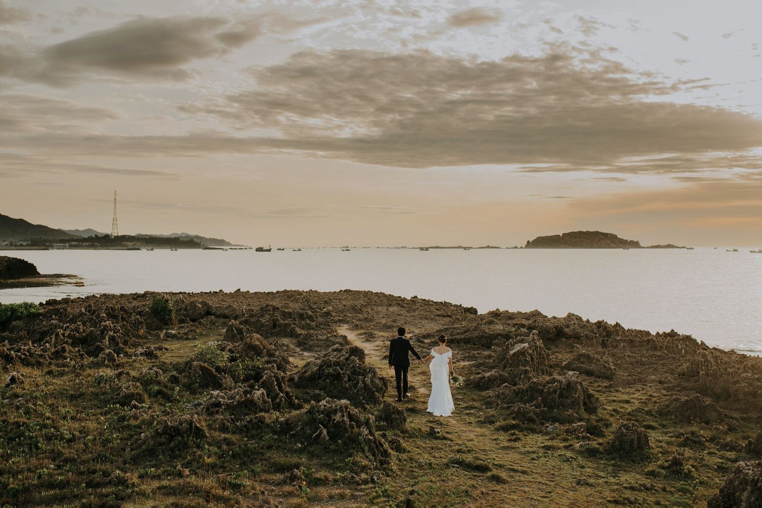 A couple stands on a coastal cliff at sunset overlooking the ocean and distant islands.