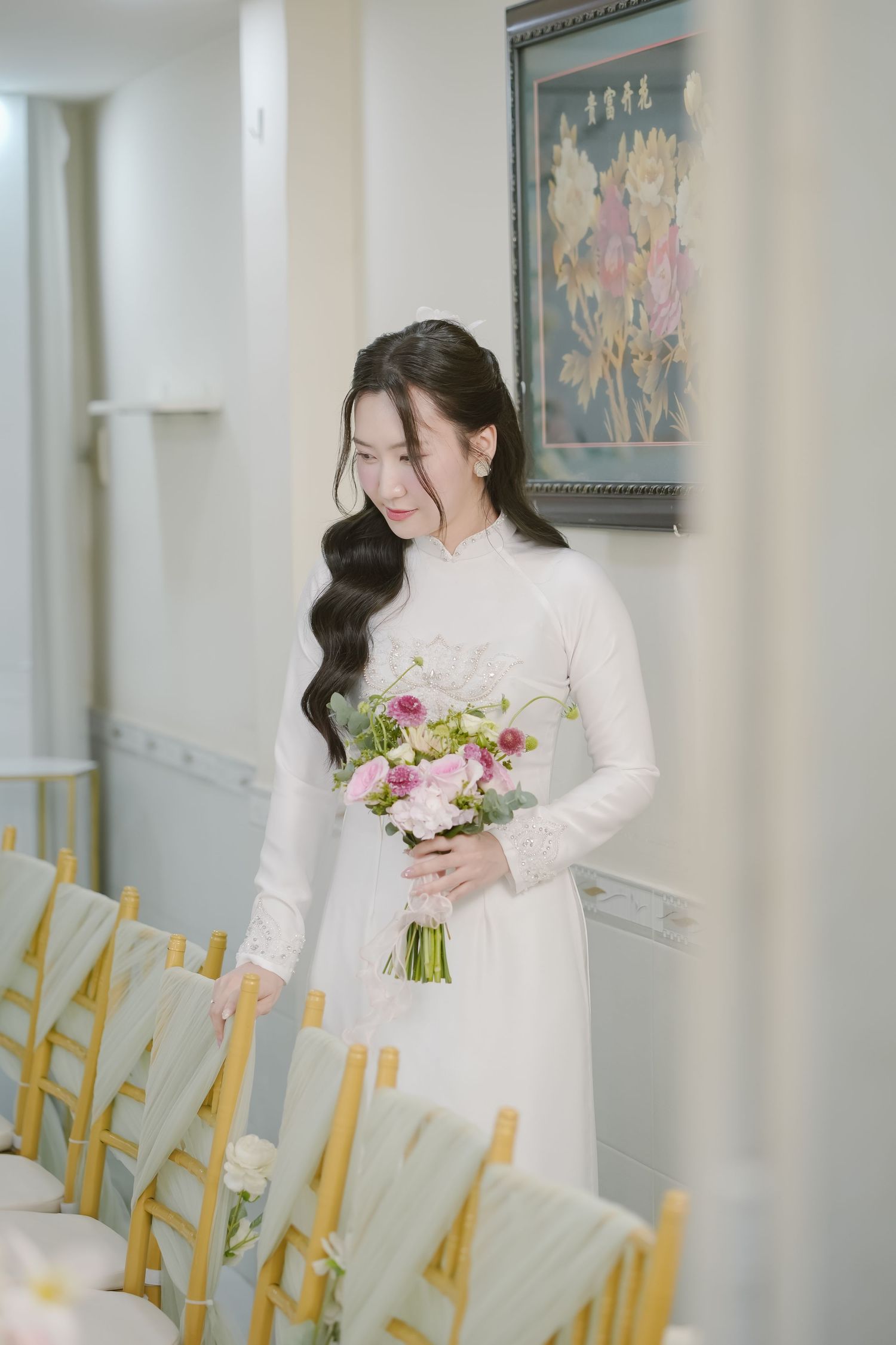 Person in traditional white ao dai holding pink floral bouquet in event space with yellow chairs.