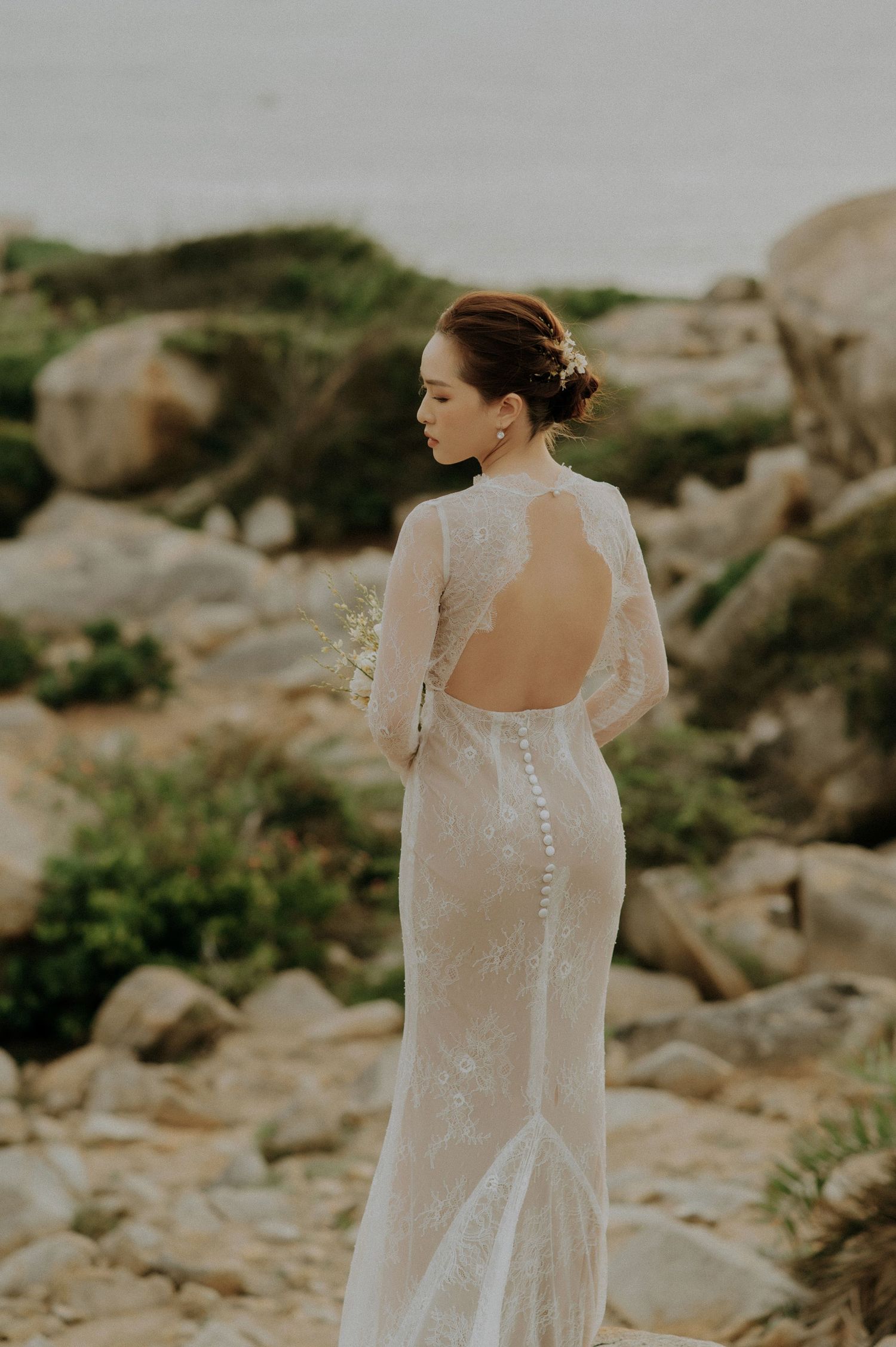 Elegant backless lace wedding dress with button details photographed outdoors against rocky natural landscape.