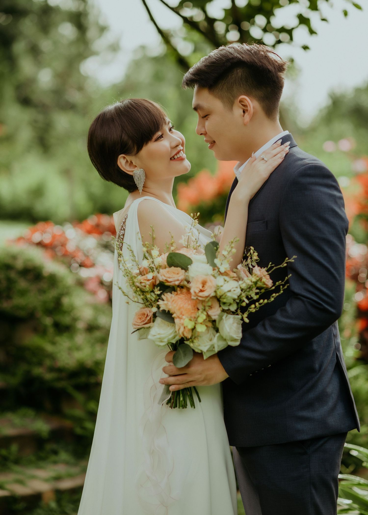 A romantic wedding couple shares an intimate moment together in a garden with peach roses and greenery.