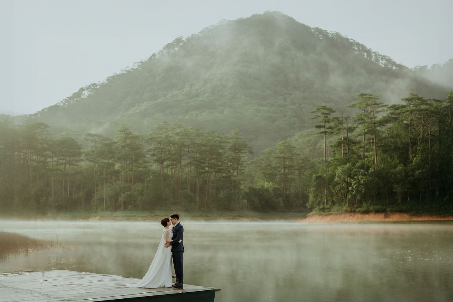A misty lake surrounded by pine trees and mountains with two figures embracing on a wooden dock.