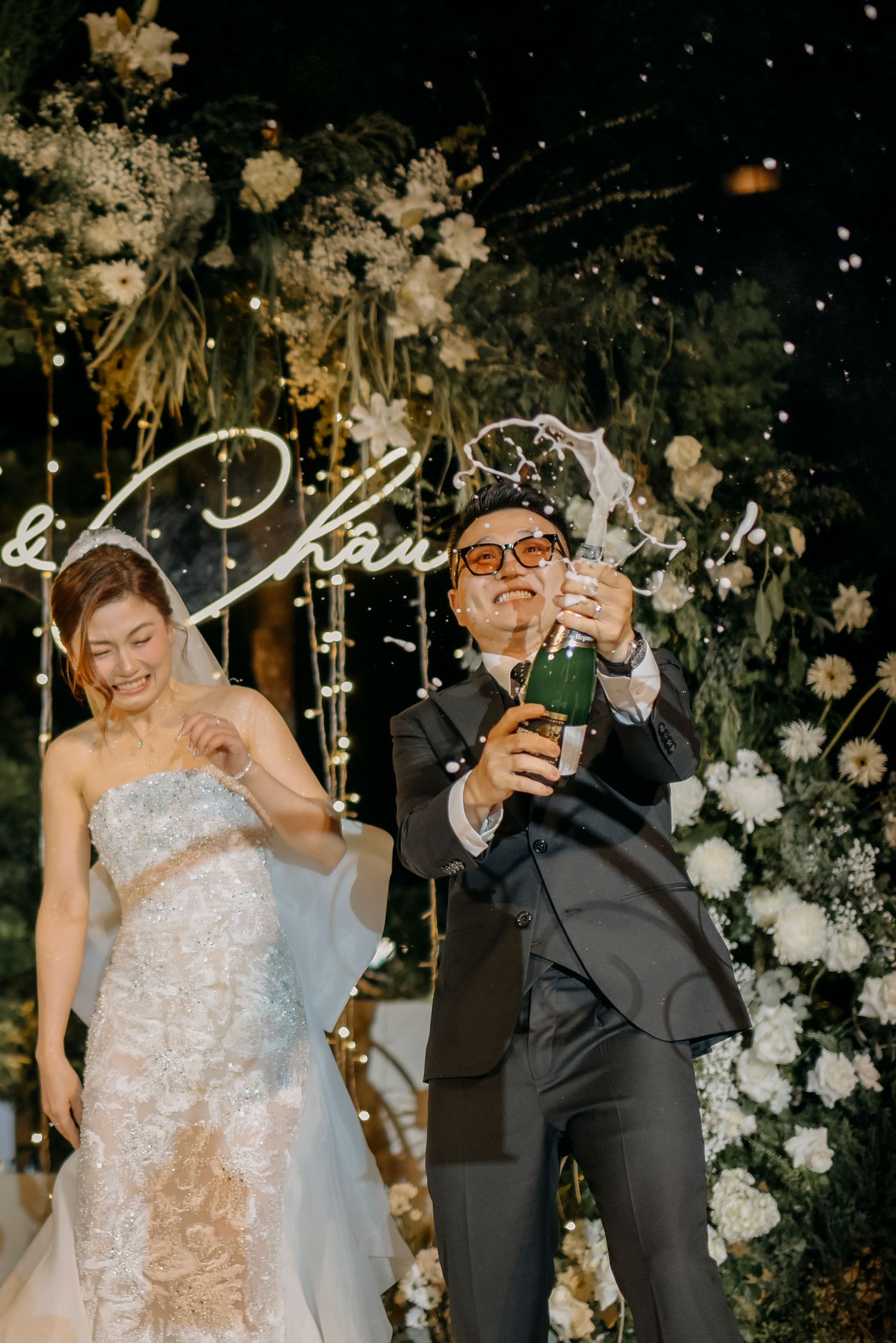 Celebratory moment with champagne during wedding reception beneath floral arch and neon signage.