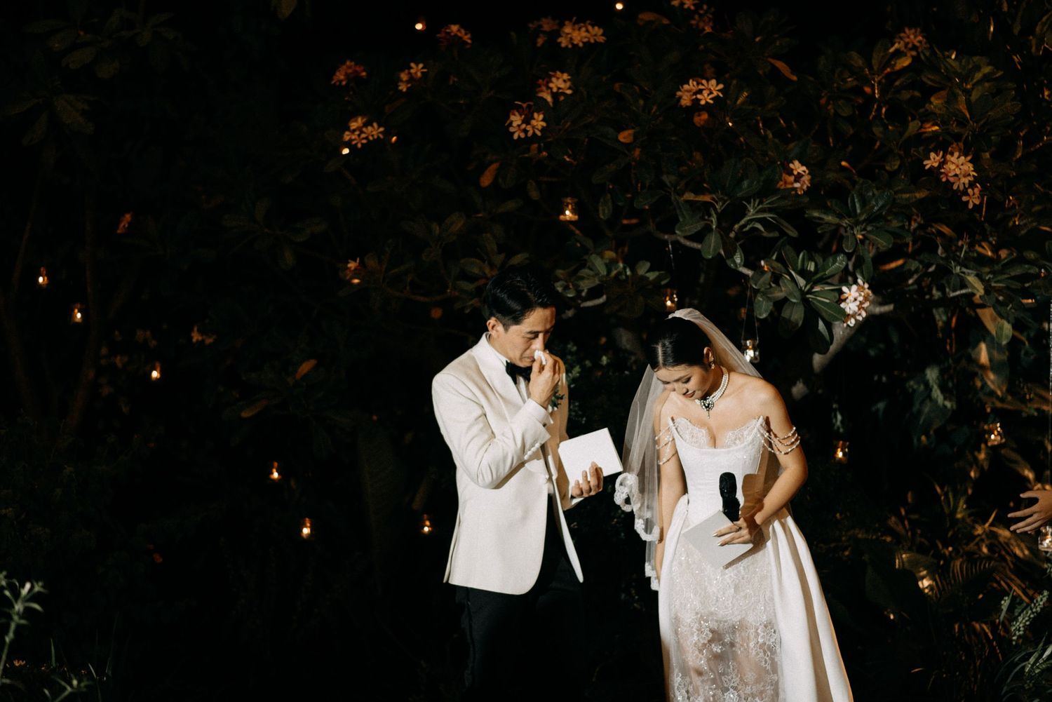 Nighttime wedding portrait of couple laughing together among illuminated foliage.