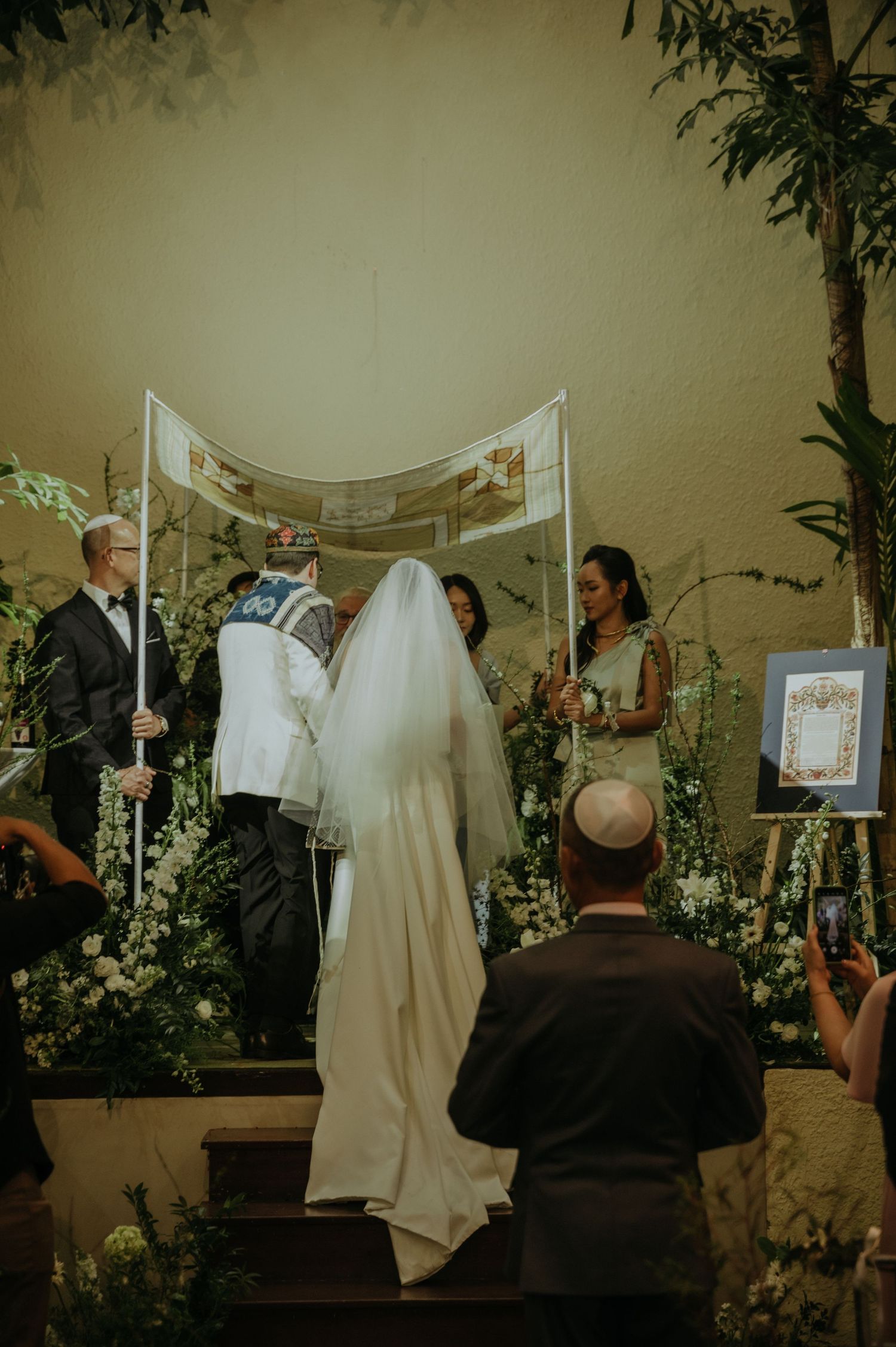 Wedding ceremony in progress under a white canopy with floral decorations and gathered guests.