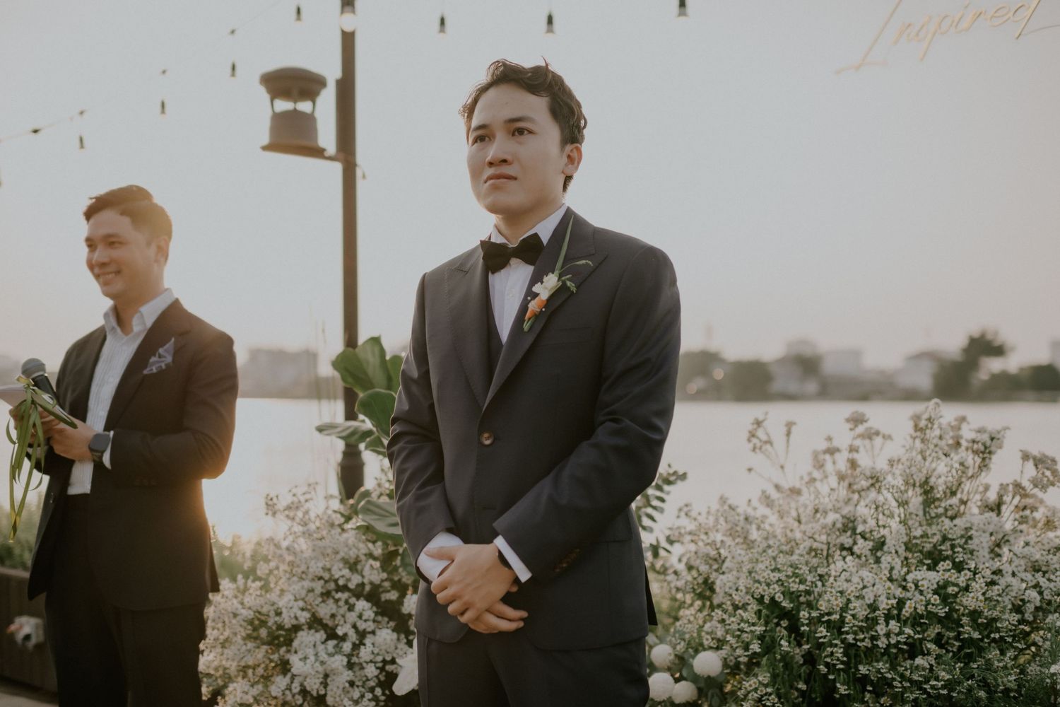 Two figures in formal attire stand outdoors against an evening sky.