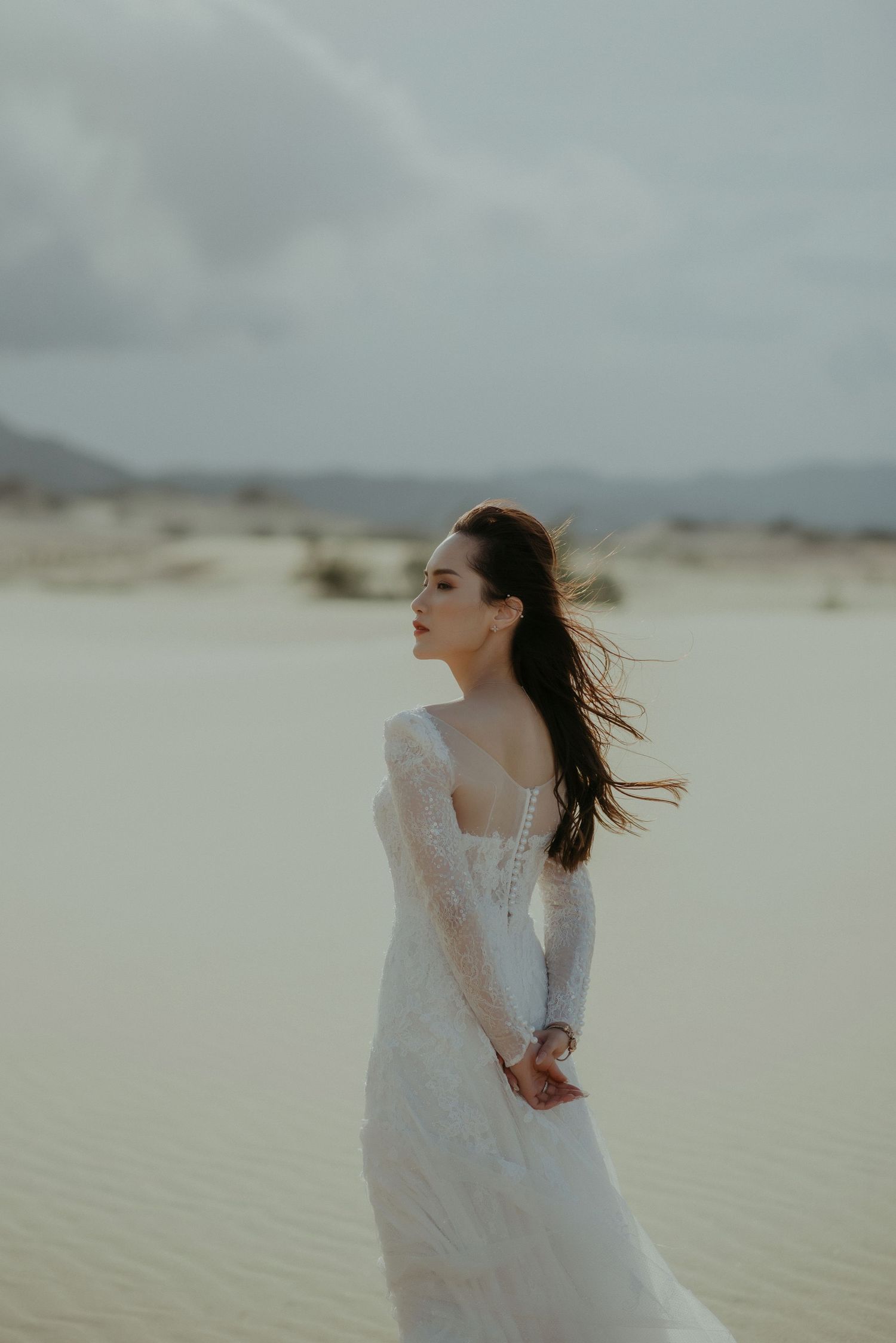 A bride in a flowing white lace dress stands gracefully in a desert landscape with windswept hair against cloudy skies.