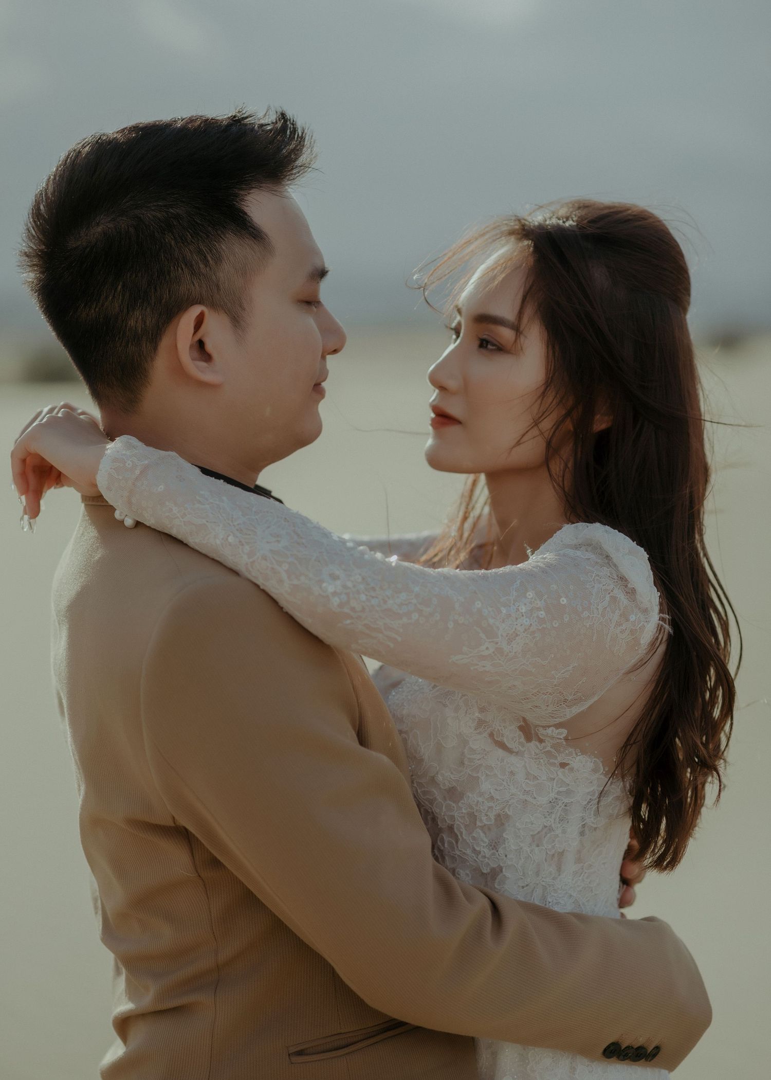 A romantic couple embraces outdoors, with the woman wearing a white lace dress and the man in a tan suit jacket.