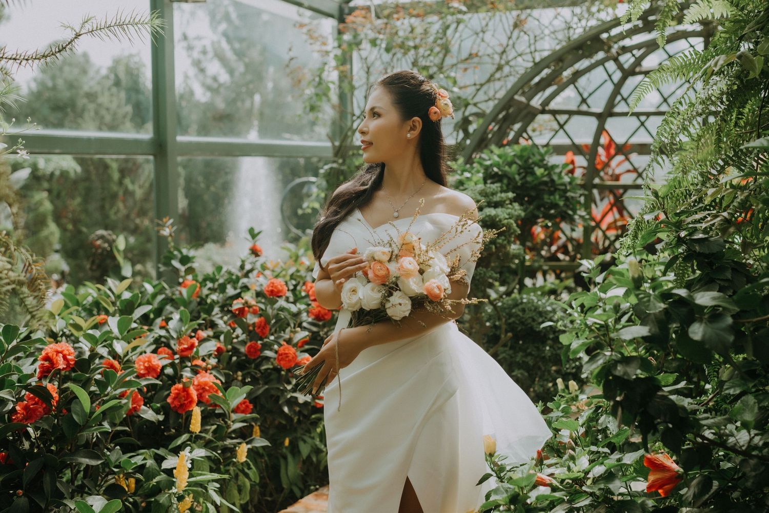 A bride in a white off-shoulder gown holds a bouquet while standing in a greenhouse filled with red flowers and greenery.