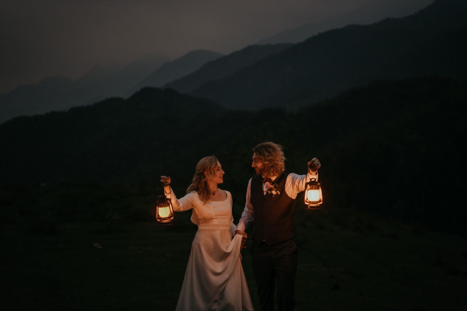 A couple holds lanterns while celebrating their wedding at dusk in the mountains.