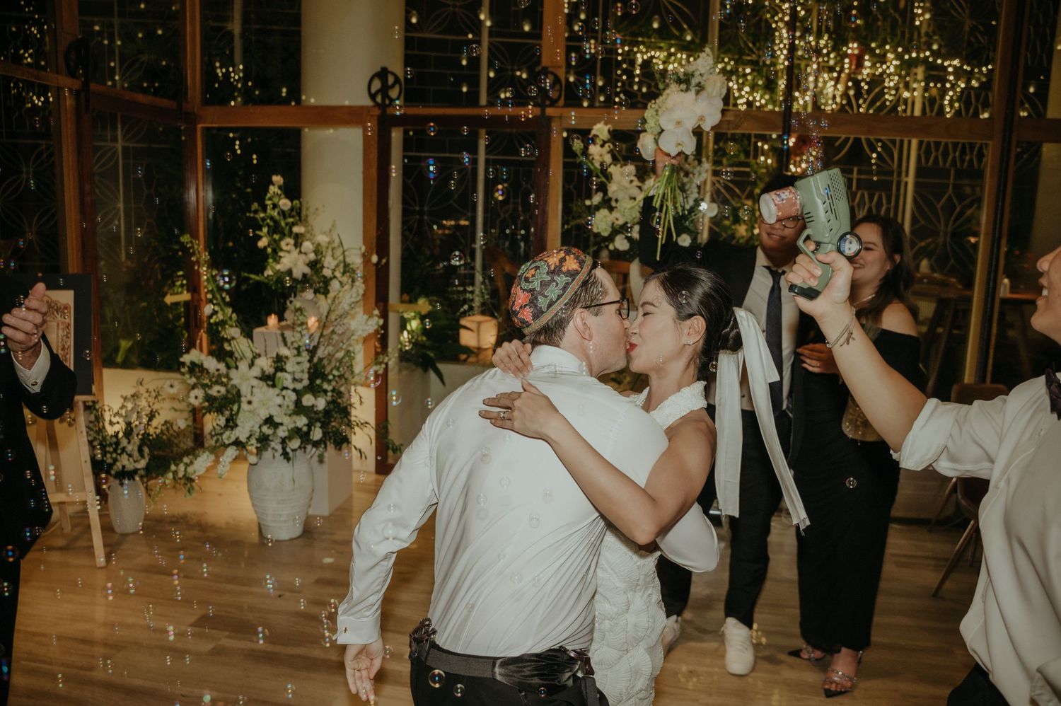 Wedding party celebrating on dance floor surrounded by floral arrangements and twinkle lights.