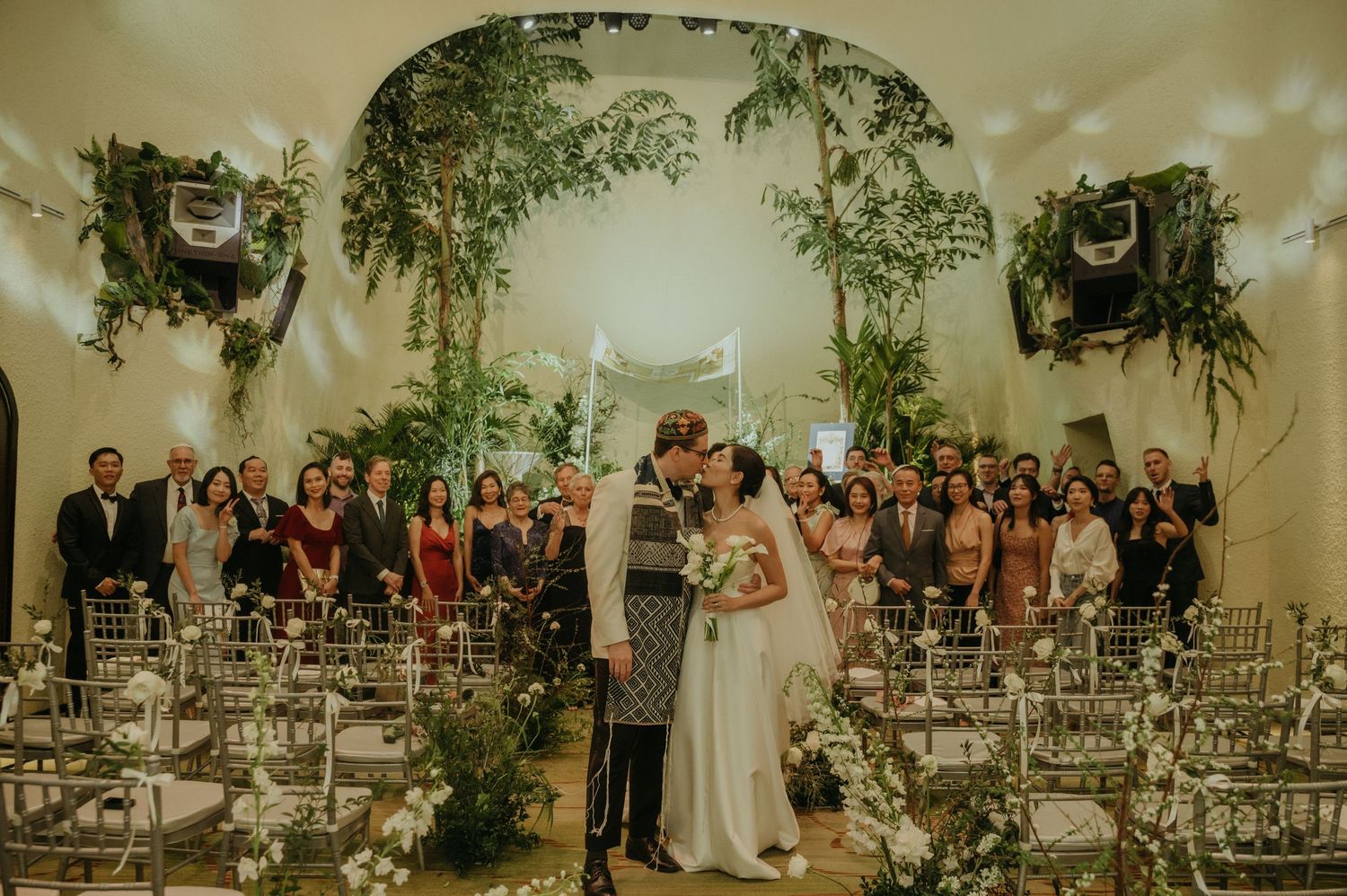 A wedding ceremony setup with white chairs and lush greenery in an arched indoor space.