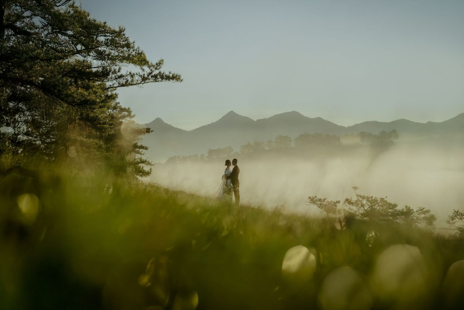 Silhouettes stand together in a misty mountain landscape at sunset with soft bokeh in the foreground.