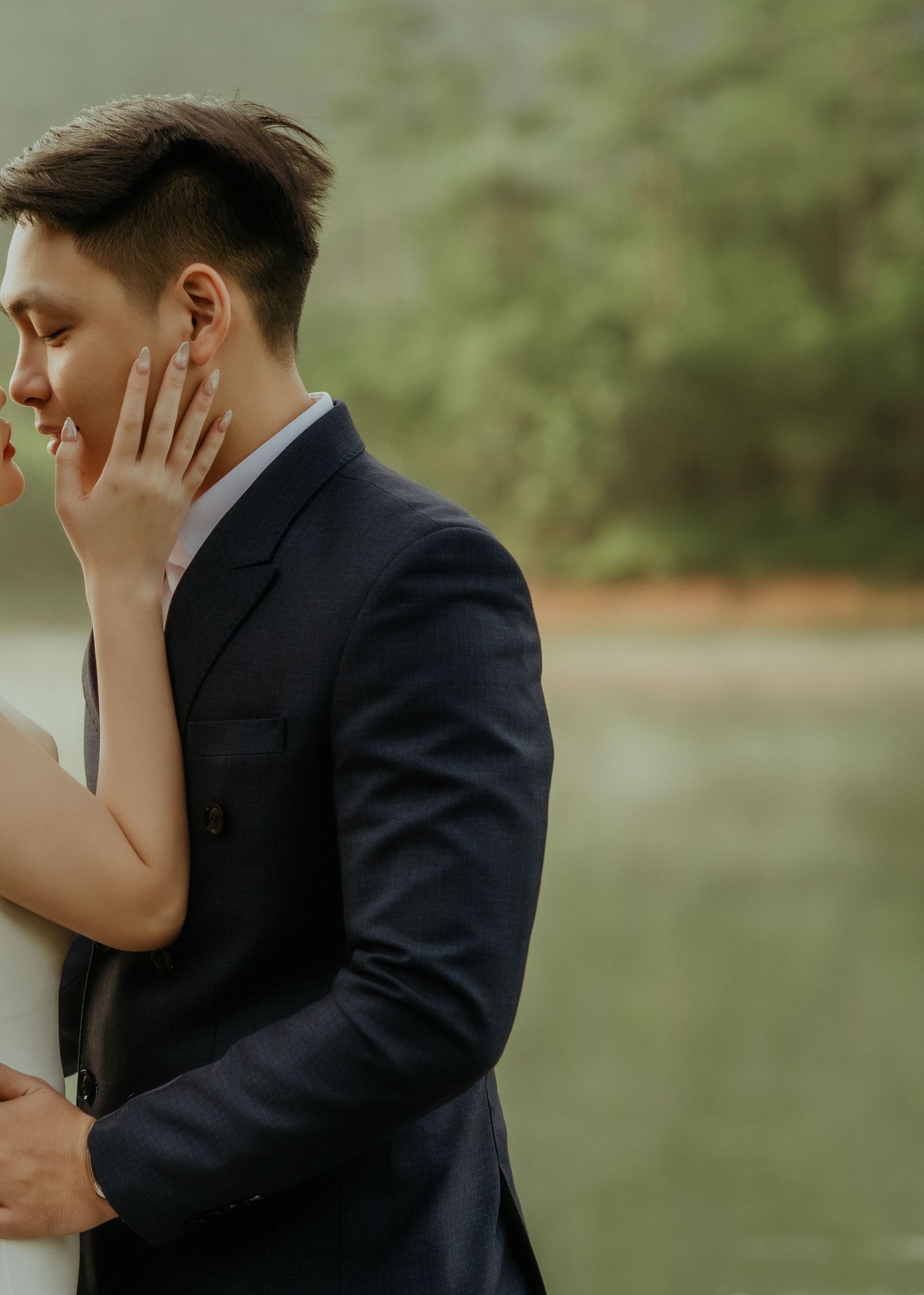 A tender moment between a couple in formal attire embracing by a peaceful lake with soft natural lighting.
