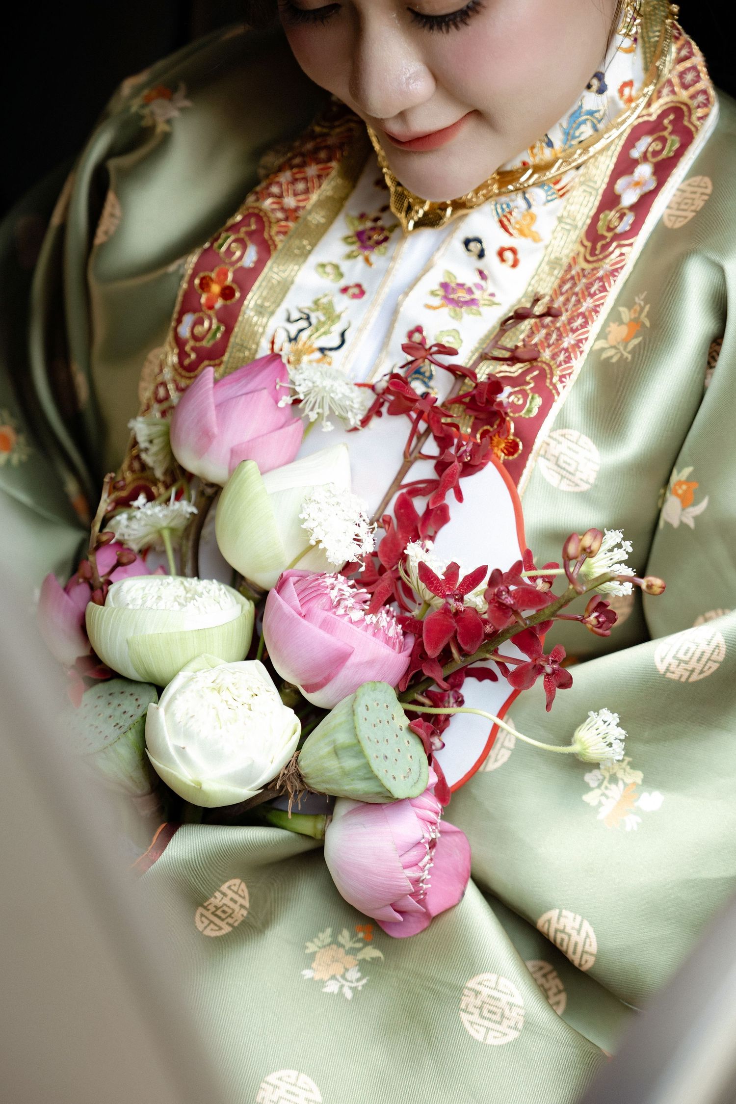 Traditional Asian bride wearing embroidered silk holds elegant bouquet of pink and white lotus buds with red orchids.