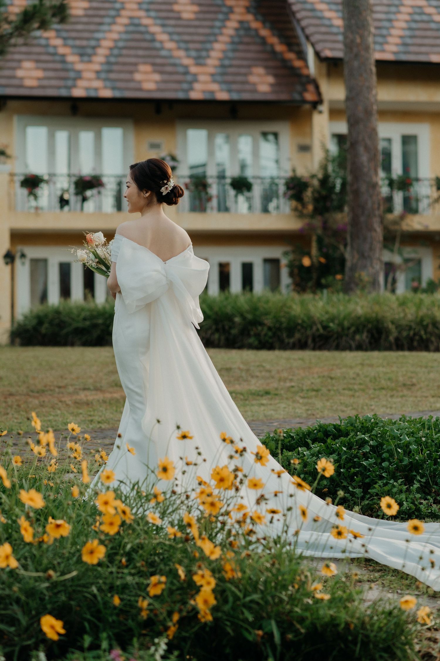 A bride in a flowing white dress stands in a garden with blooming yellow flowers against a rustic building backdrop.
