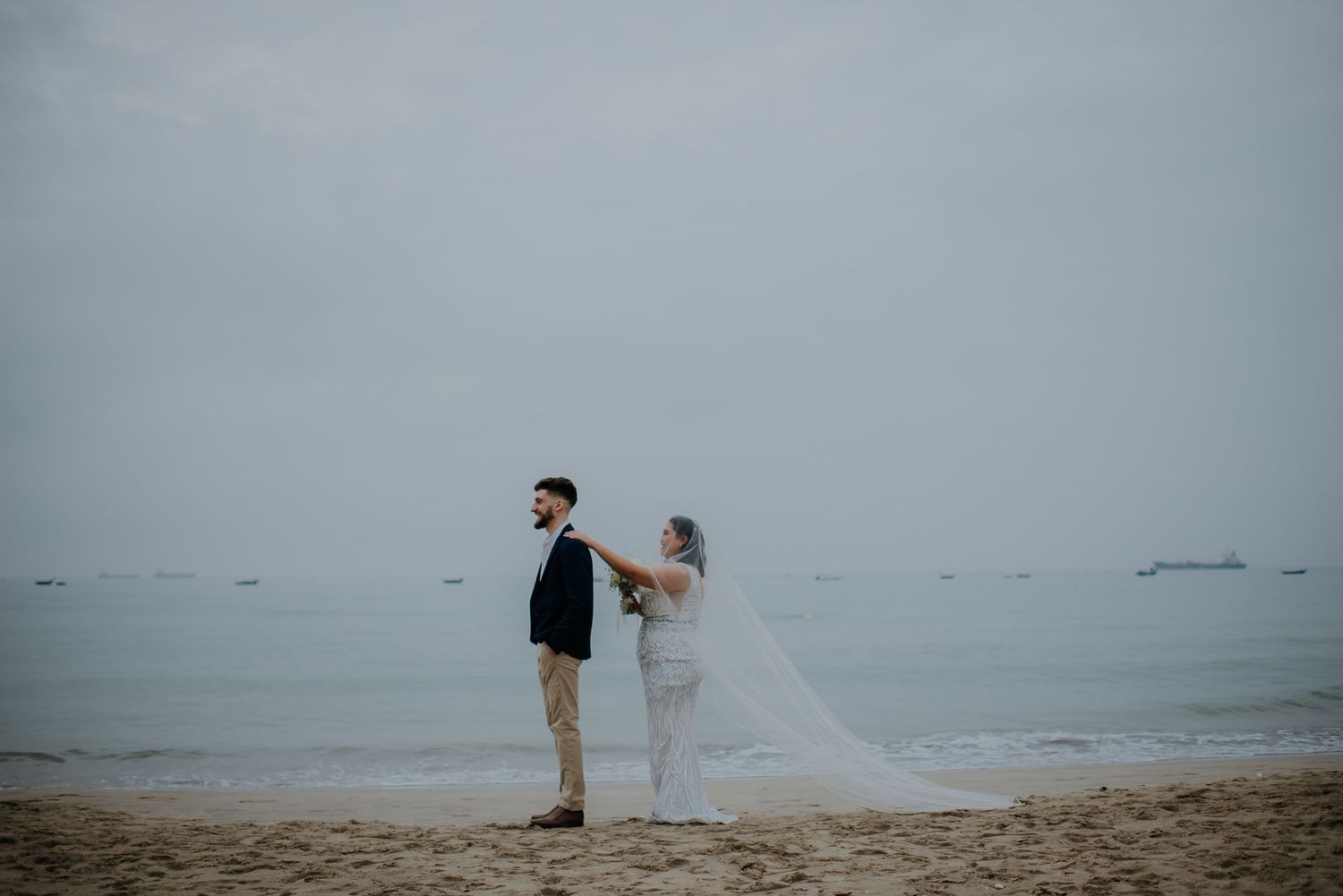 Dramatic wedding photo of couple standing on misty beach with cityscape in background.