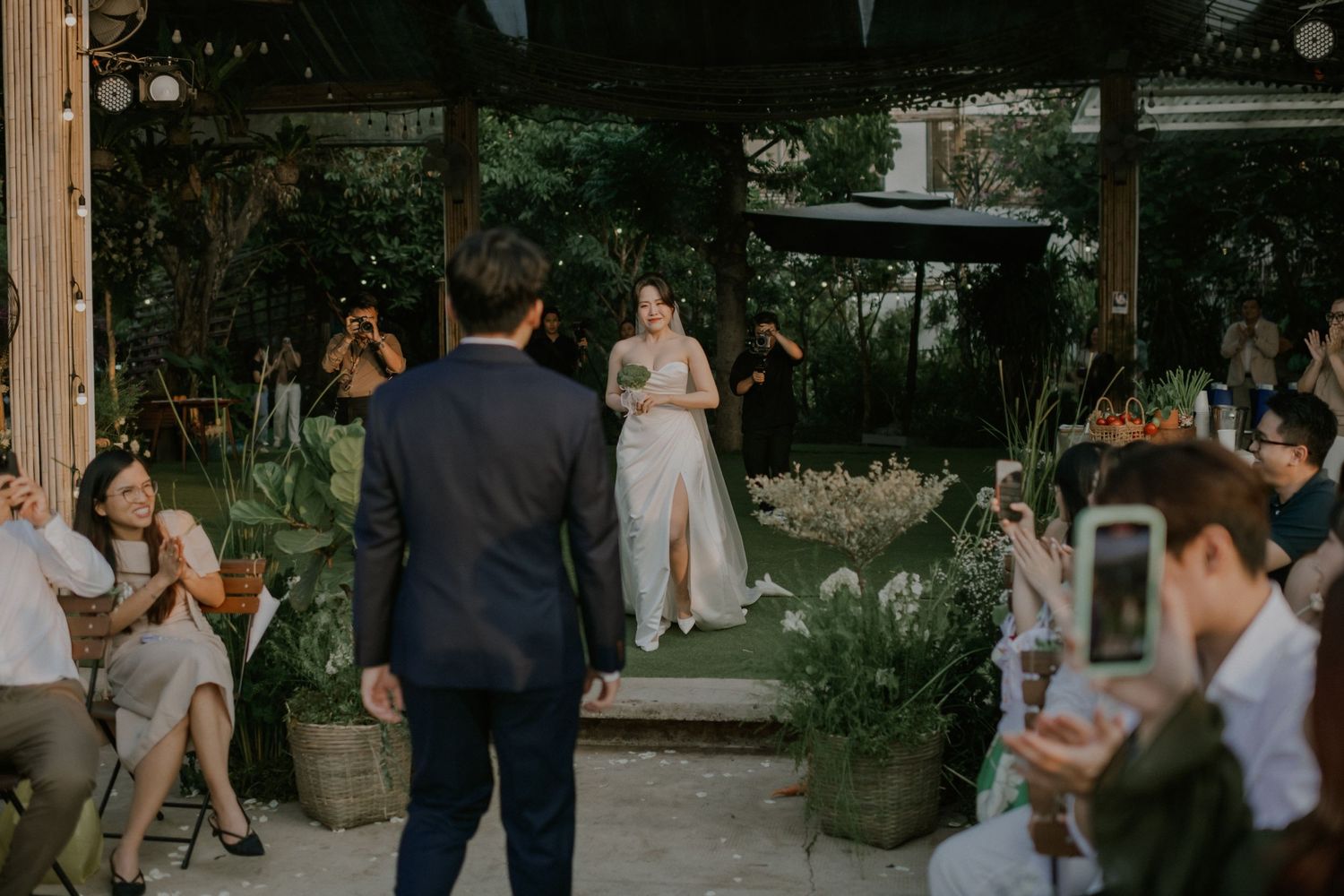 Wedding guests watch a ceremony in a garden setting with string lights overhead.
