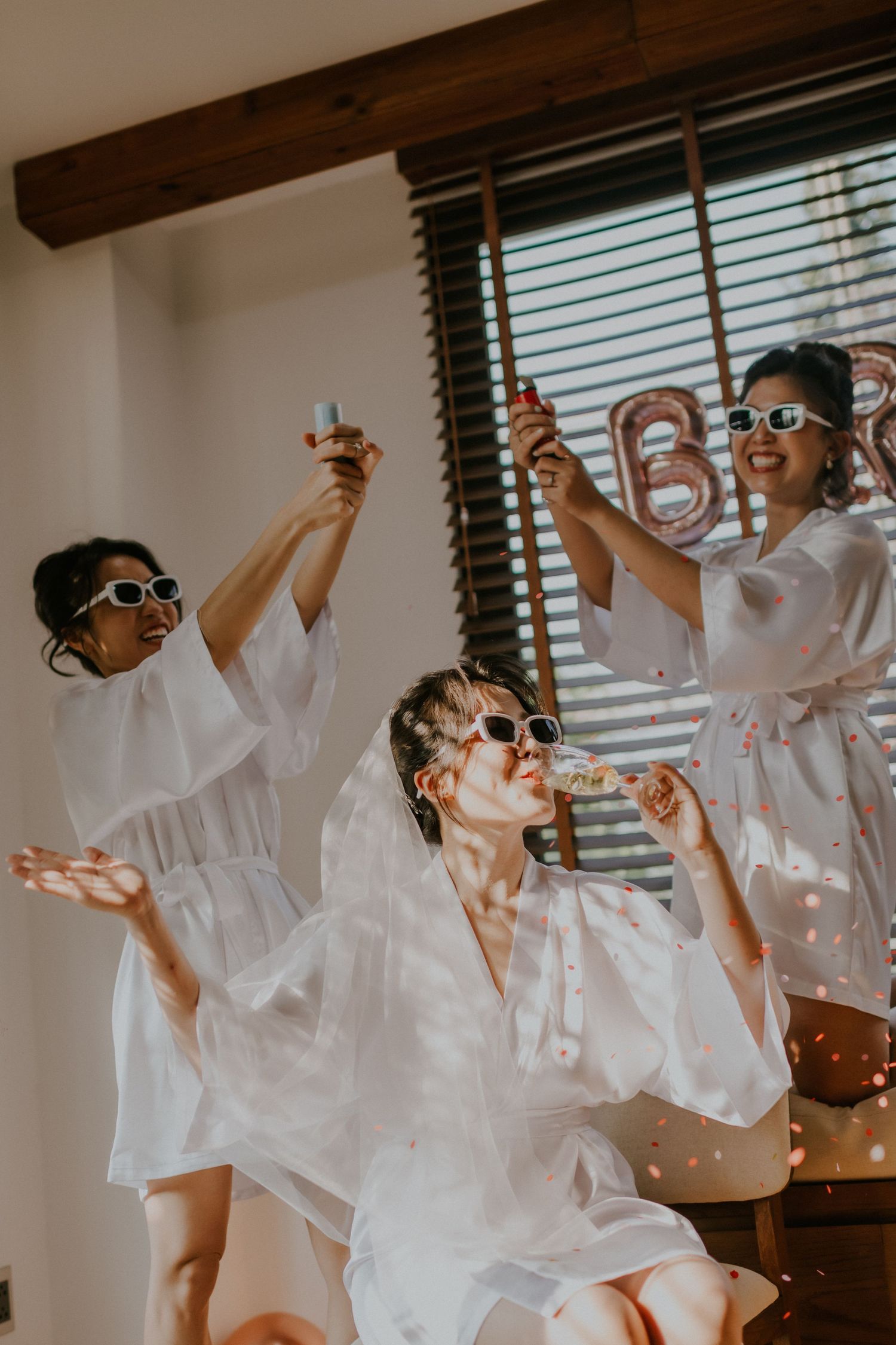 Wedding party in white robes and sunglasses pose playfully while holding drinks.