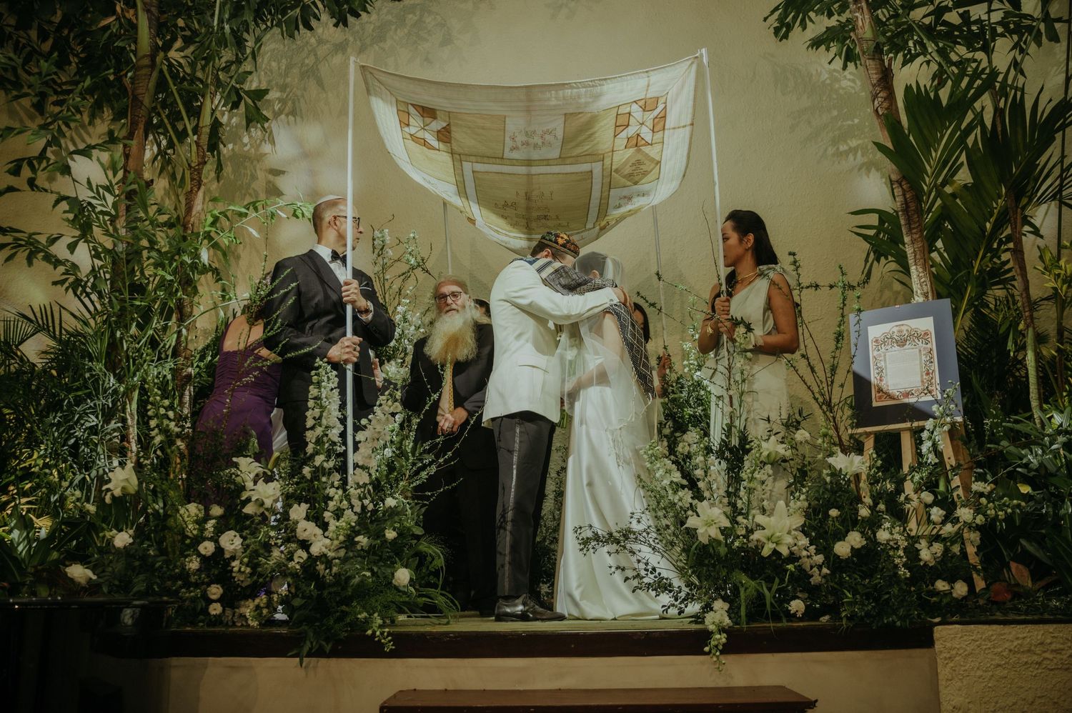 A wedding ceremony on a raised platform surrounded by lush floral arrangements and hanging fabric.