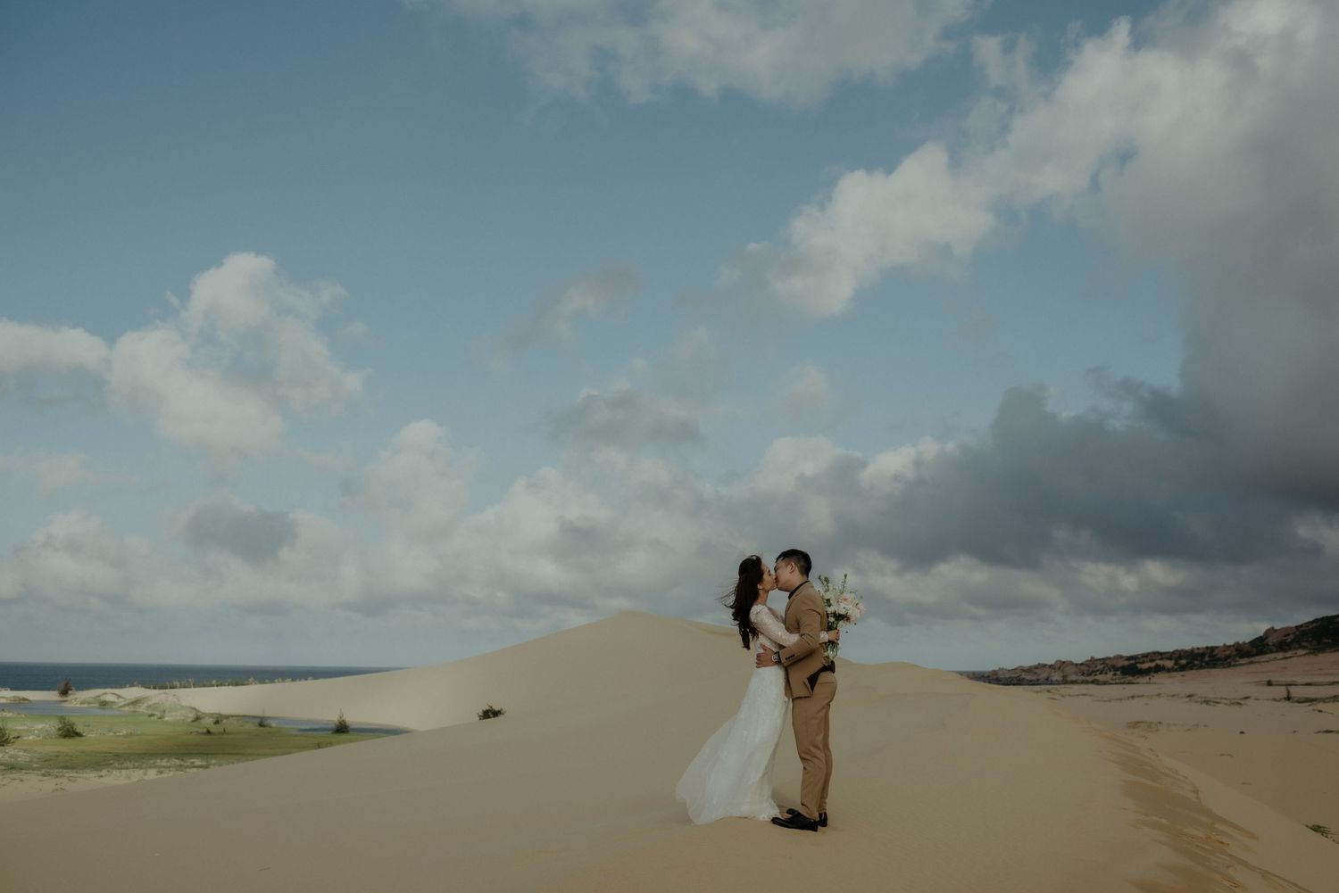 Couple embraces on a scenic sandy beach dune under cloudy blue skies.