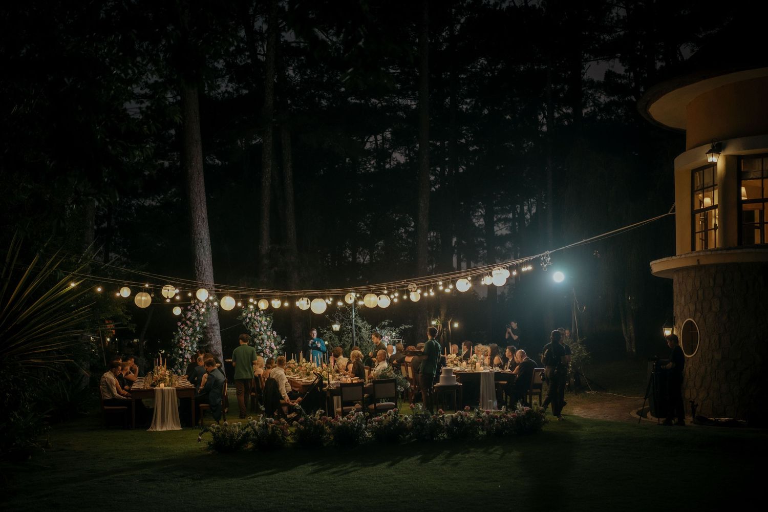 An outdoor wedding reception at night with string lights illuminating guests dining at tables under dark trees.
