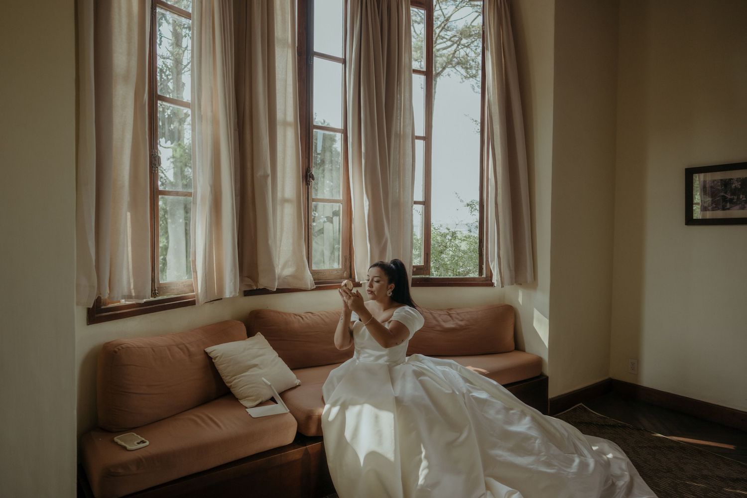 A person in a white wedding dress sits on a tan leather couch by large windows in soft natural light.