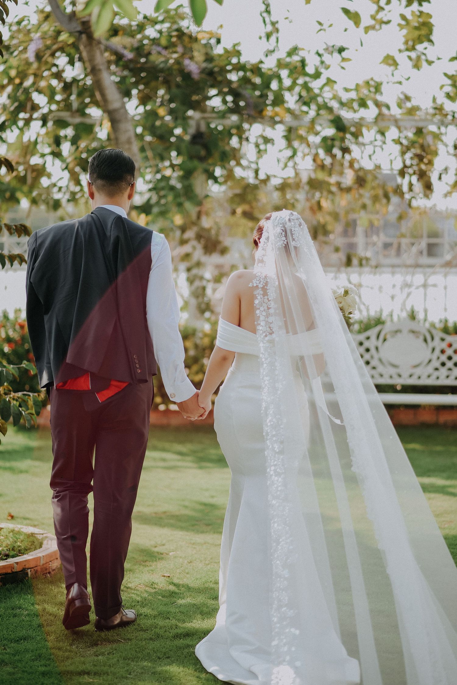 Newlyweds walk hand in hand through a garden setting with the bride wearing a flowing veil and fitted white dress.