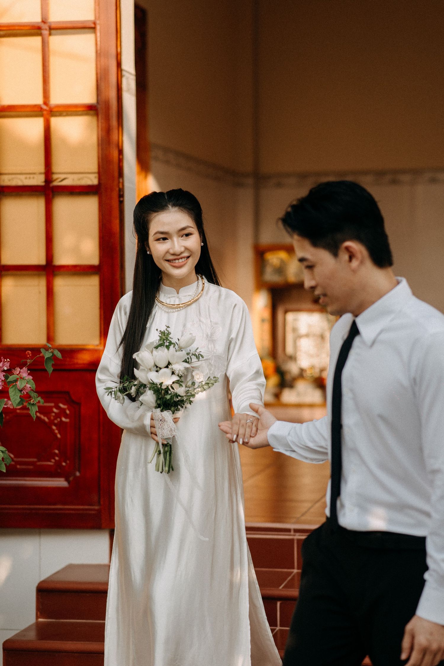 Two people in formal attire stand together on red steps holding bouquet of white flowers.