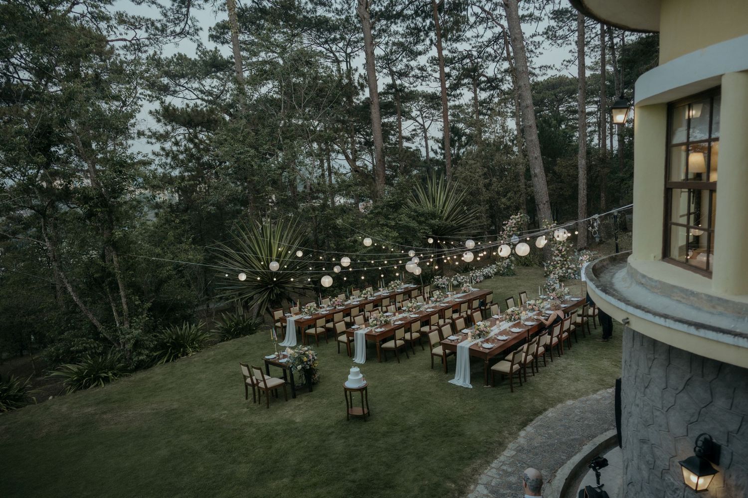 Aerial view of an elegant outdoor wedding reception setup with long wooden tables surrounded by pine trees at dusk.