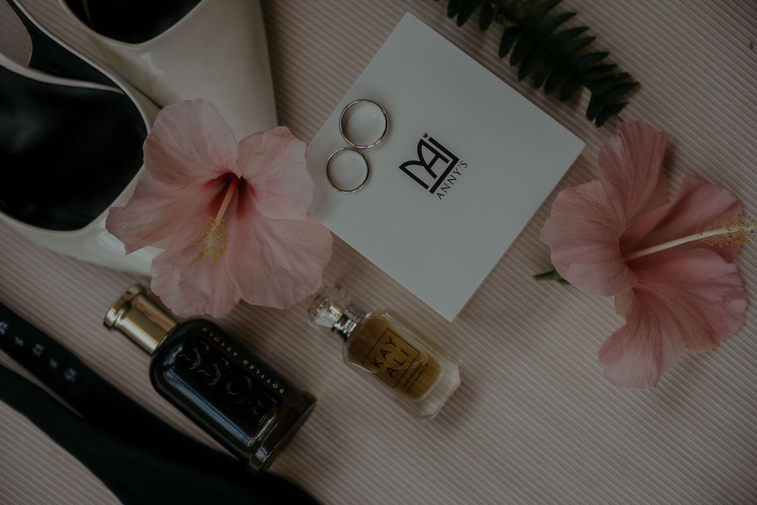 Wedding ring details with pink hibiscus flowers, perfumes and invitation card on a textured white surface.