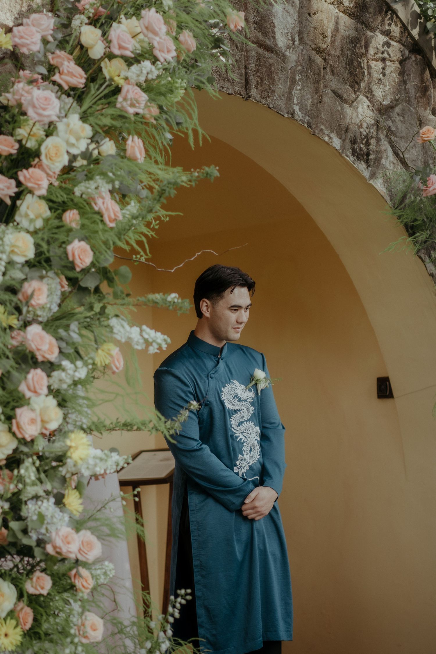 Someone wearing a traditional teal blue Ao Dai stands beneath an arched doorway adorned with pink roses and greenery.
