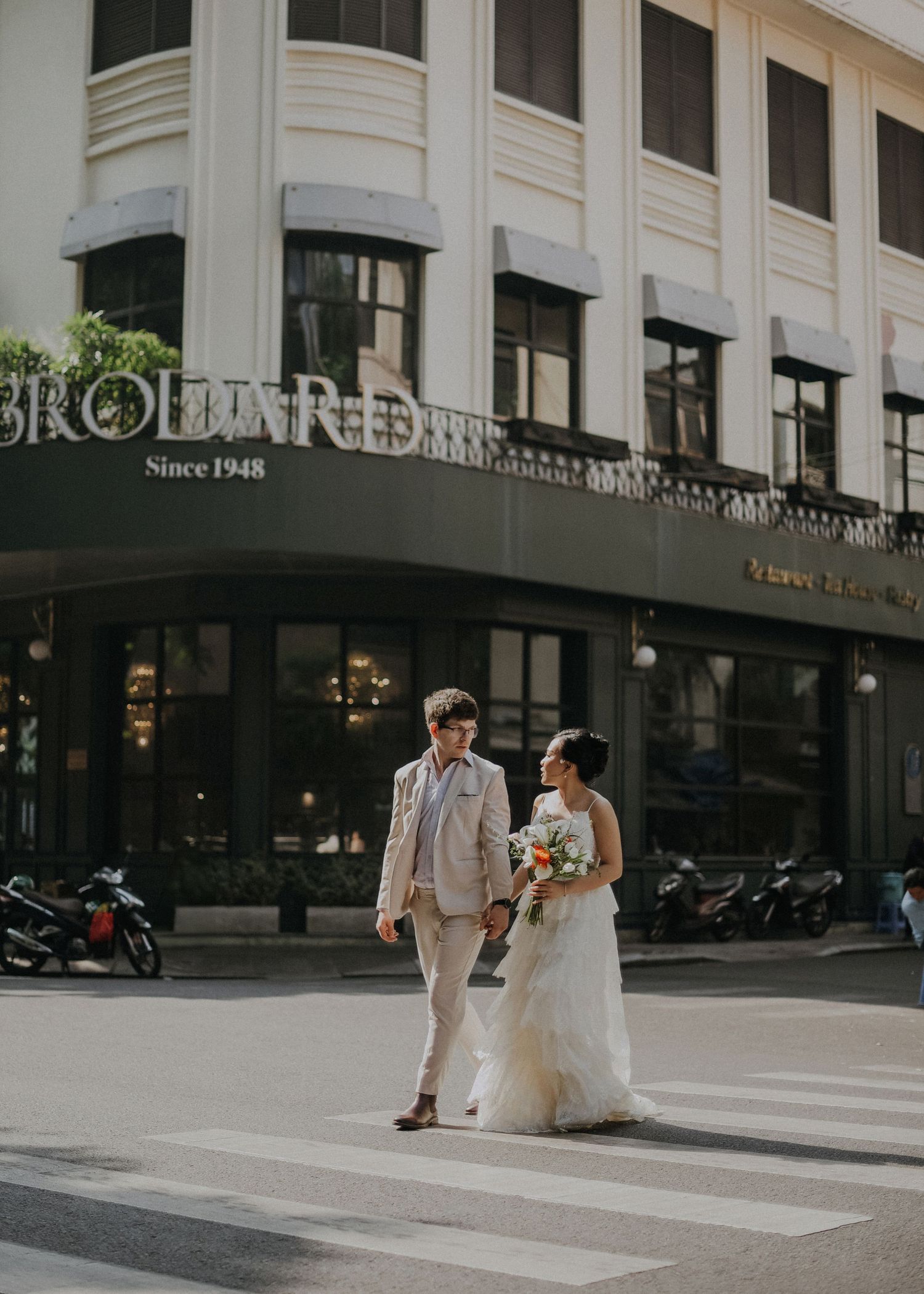 A couple walk across a crosswalk in front of a historic building storefront during their wedding photo session.