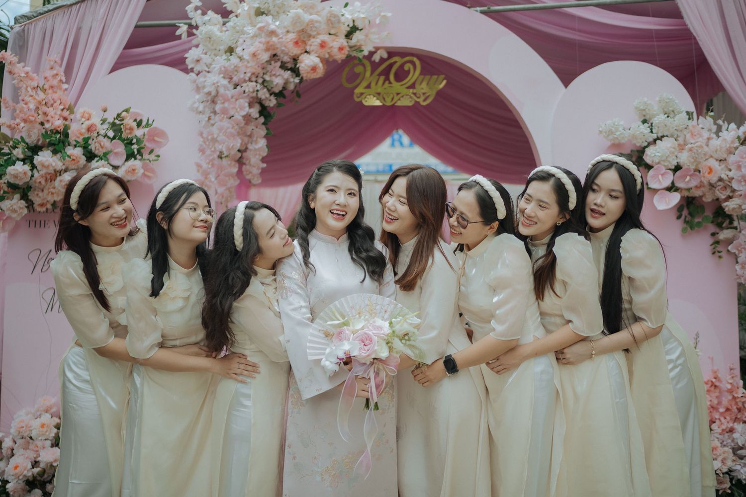 Group wearing matching cream-colored dresses poses under a pink floral arch for a formal event.