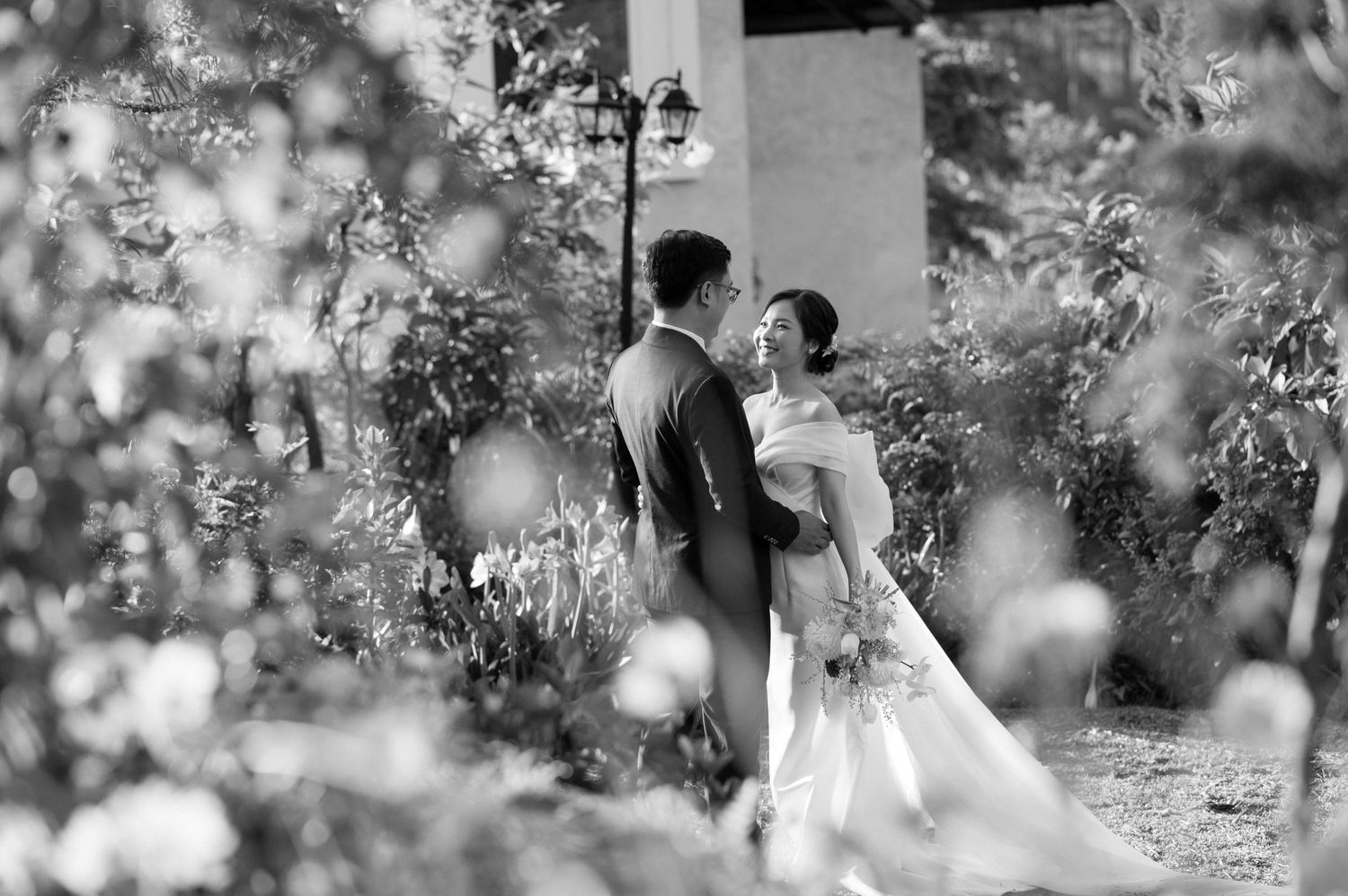 Black and white wedding photograph of a romantic couple embracing among blossoming trees and flowers in a garden setting.