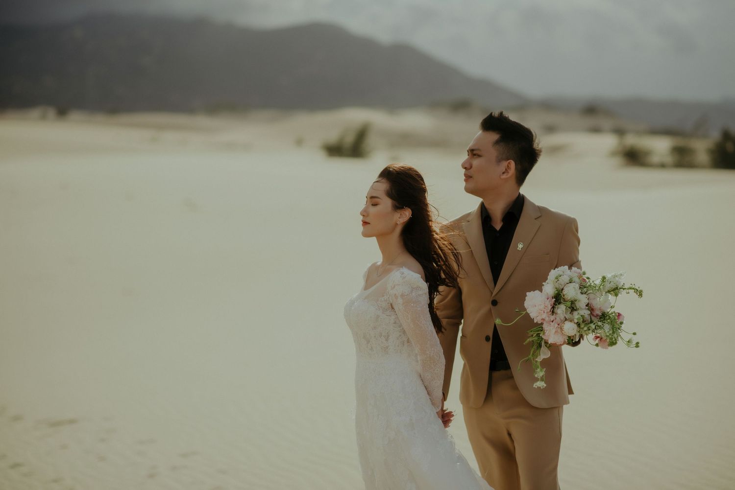 Romantic couple in bridal attire share a tender moment on a desert landscape with white flowers at sunset.