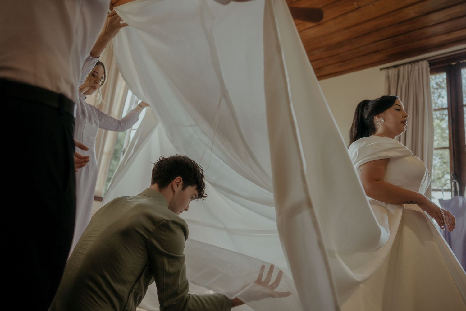 A person adjusts the flowing white wedding dress train while bride stands in elegant off-shoulder gown.
