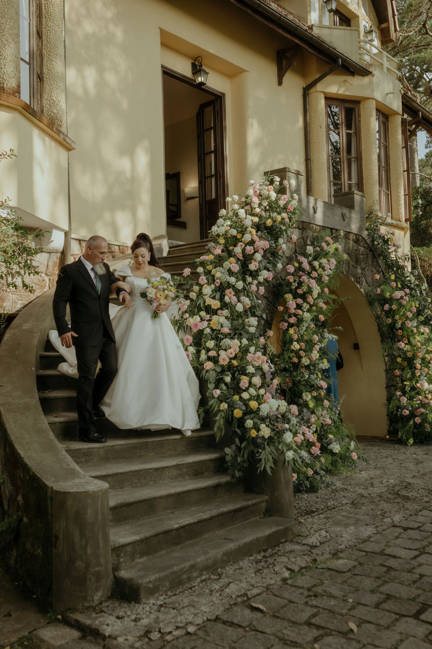 Couple standing on stone steps of vintage building with elegant floral arch displaying peach and cream roses.