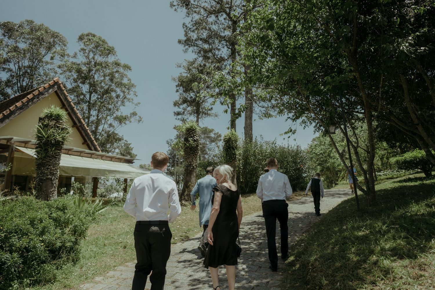 Small group walks along pathway outdoors with lush greenery and rustic architecture on a sunny day.