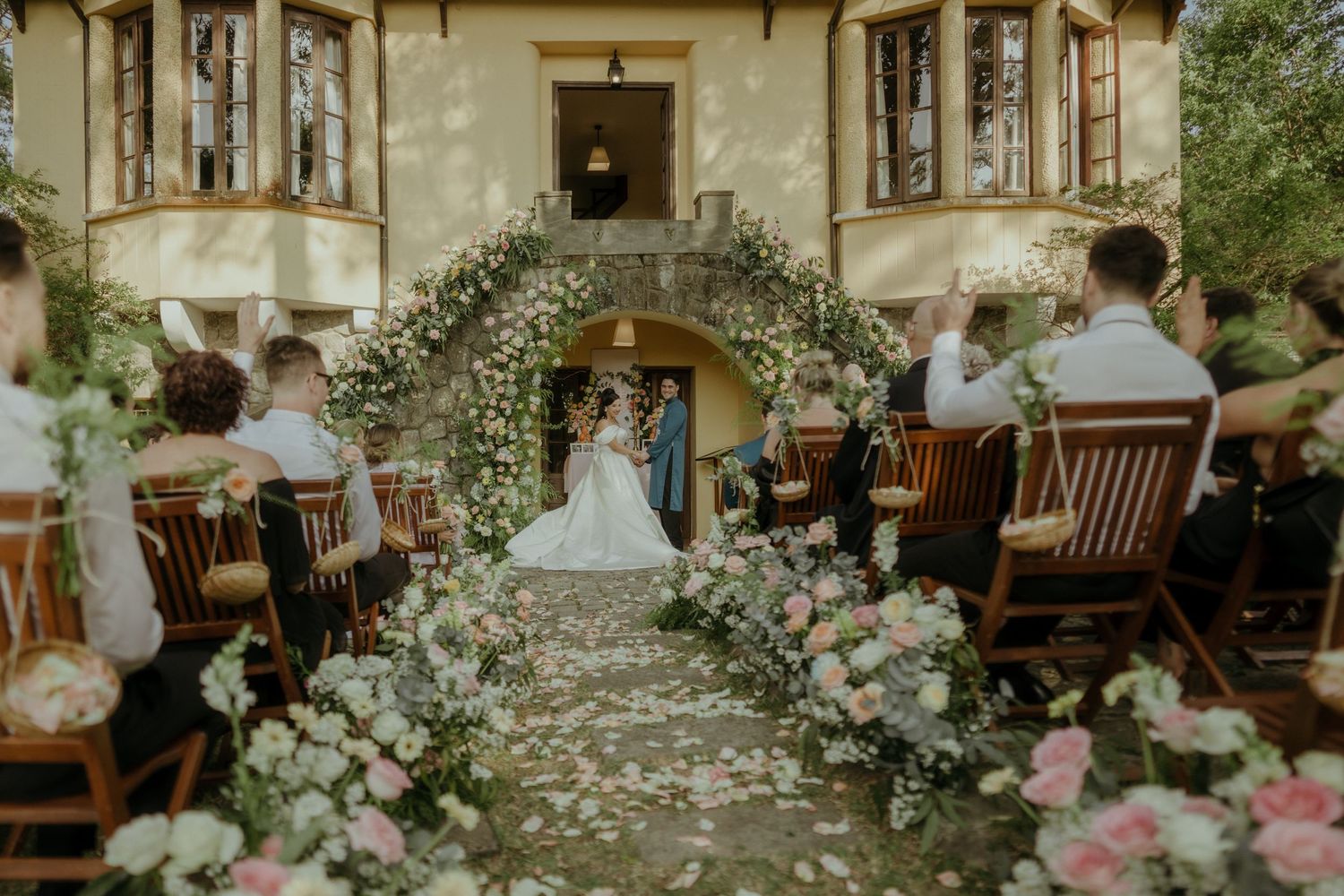 A wedding ceremony under a floral arch in a garden setting with guests seated on wooden chairs and scattered petals on the path.