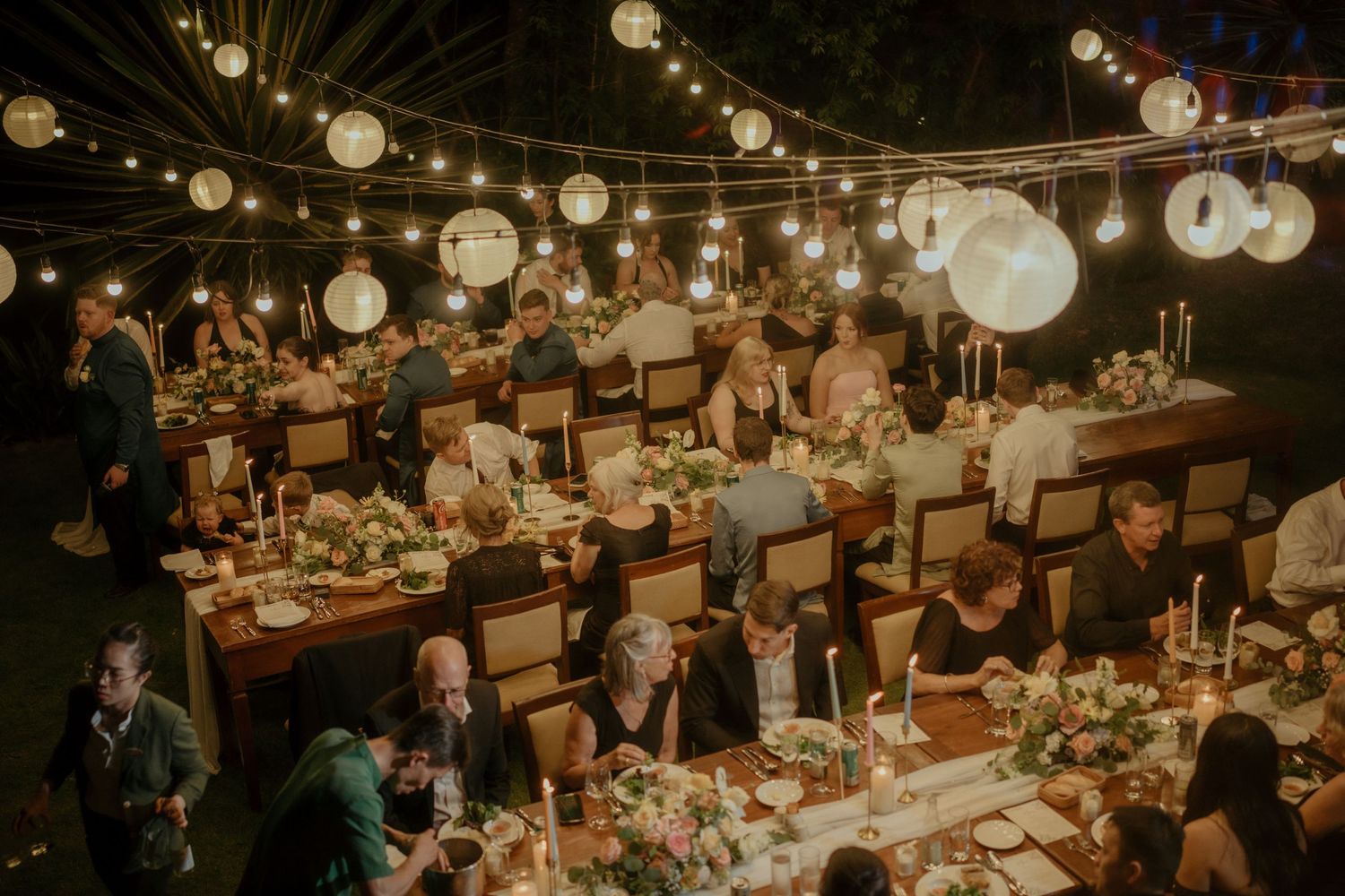 Elegant wedding reception venue with hanging paper lanterns illuminating long banquet tables set for dinner.
