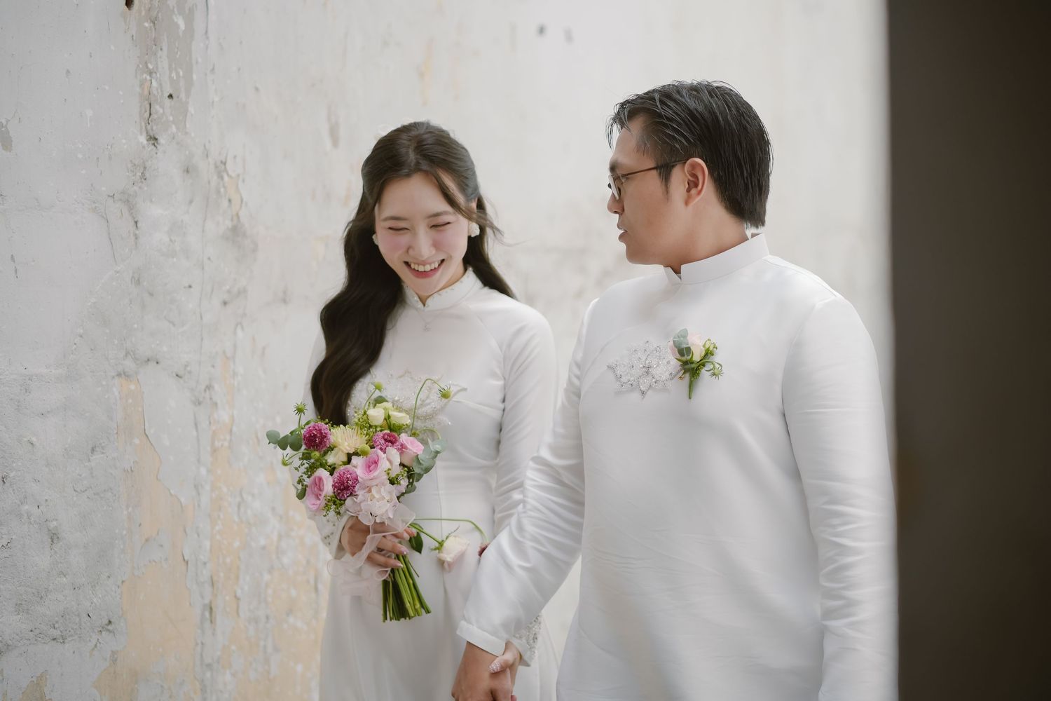Couple in white traditional attire holding pink floral bouquet against white wall.