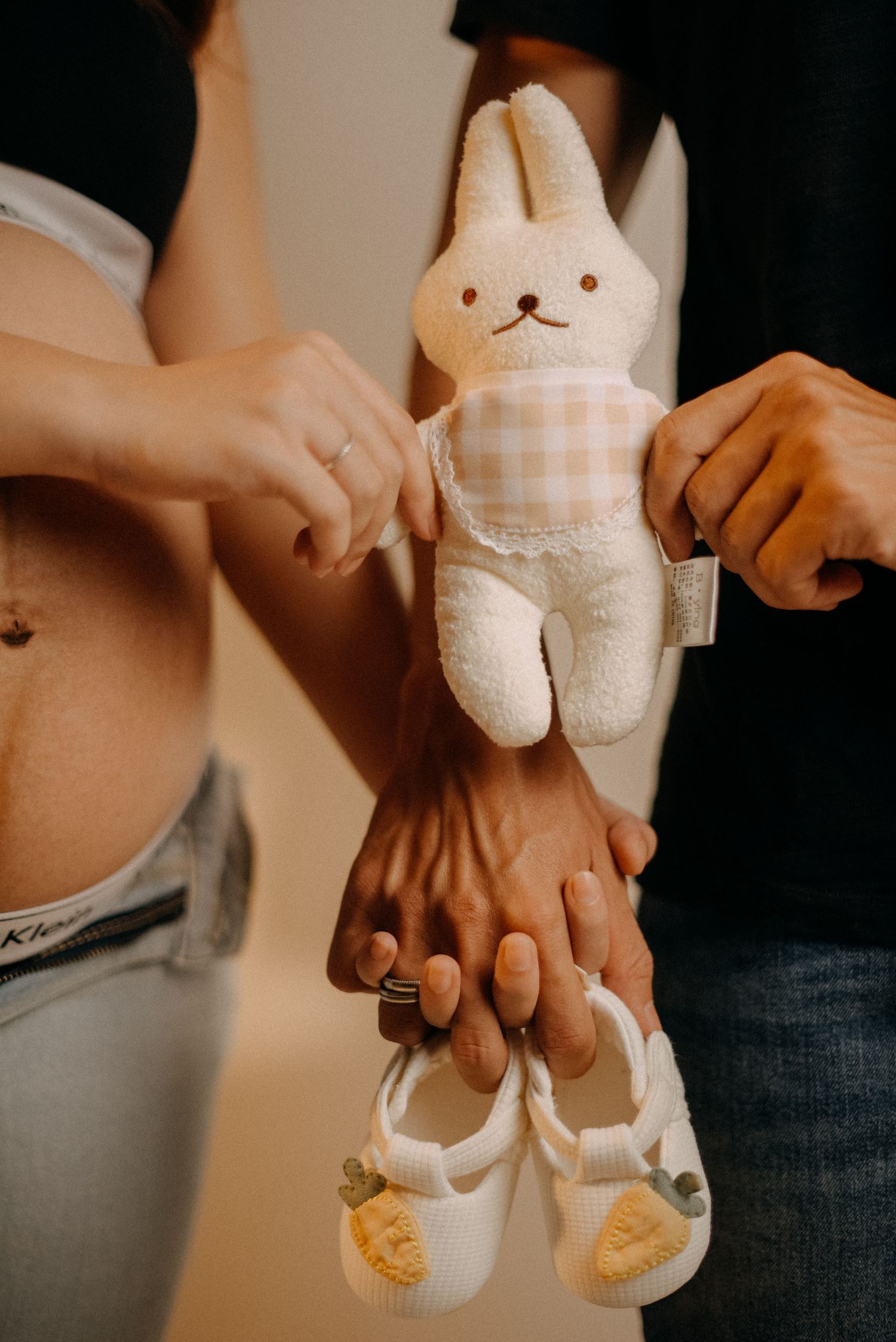 A plush bunny toy and baby shoes displayed against a pregnant belly in warm lighting.