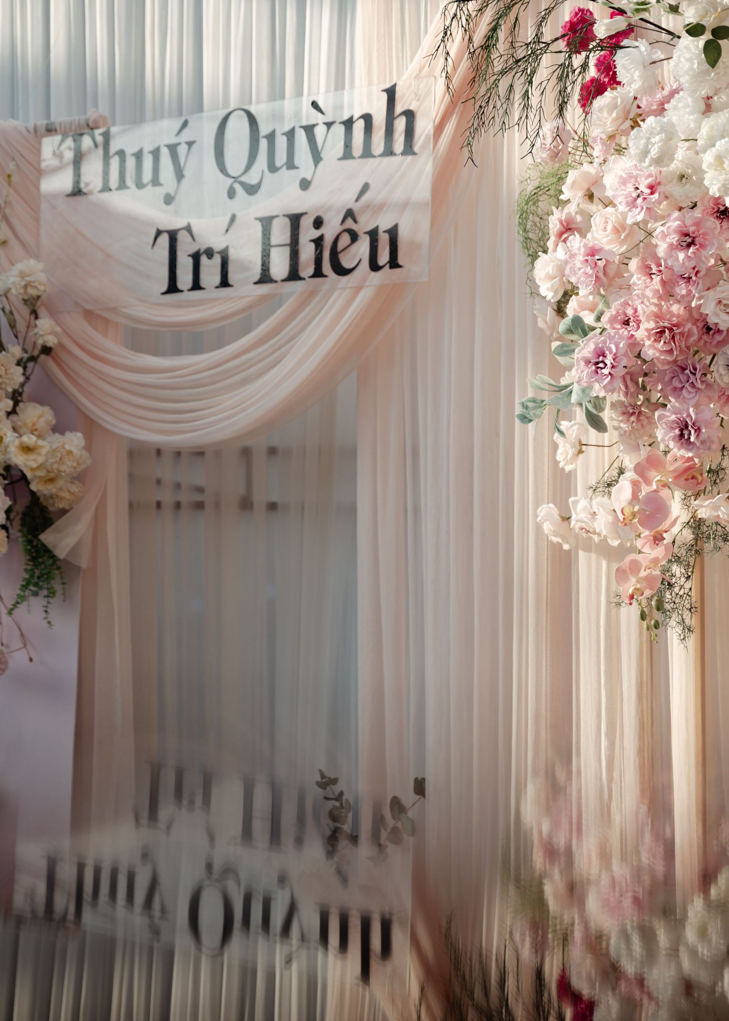 Elegant wedding backdrop with pink floral arrangements and white draping featuring Vietnamese text 'Thuy Quynh Tri Hieu'.