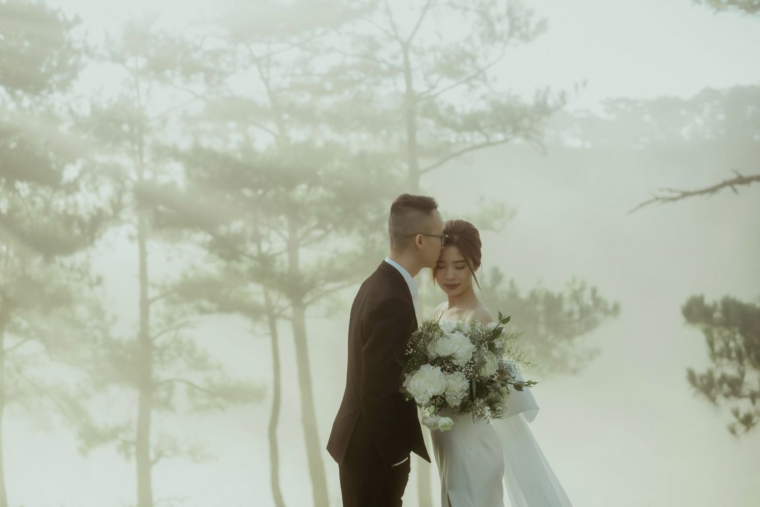 A couple in wedding attire share a tender moment in a misty forest setting with pine trees in the background.
