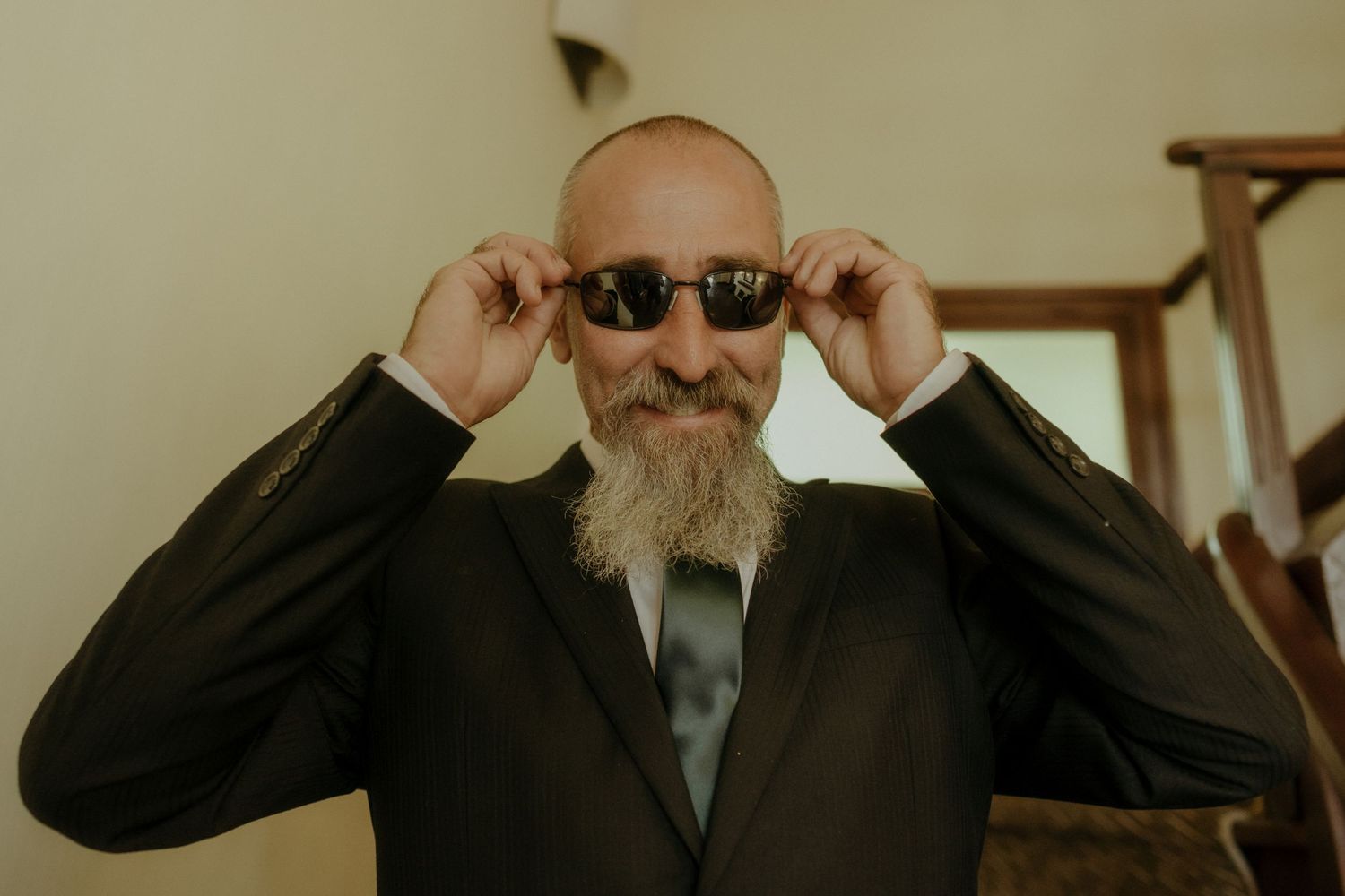 A bearded man in a dark suit adjusts his sunglasses while getting ready for a formal event.
