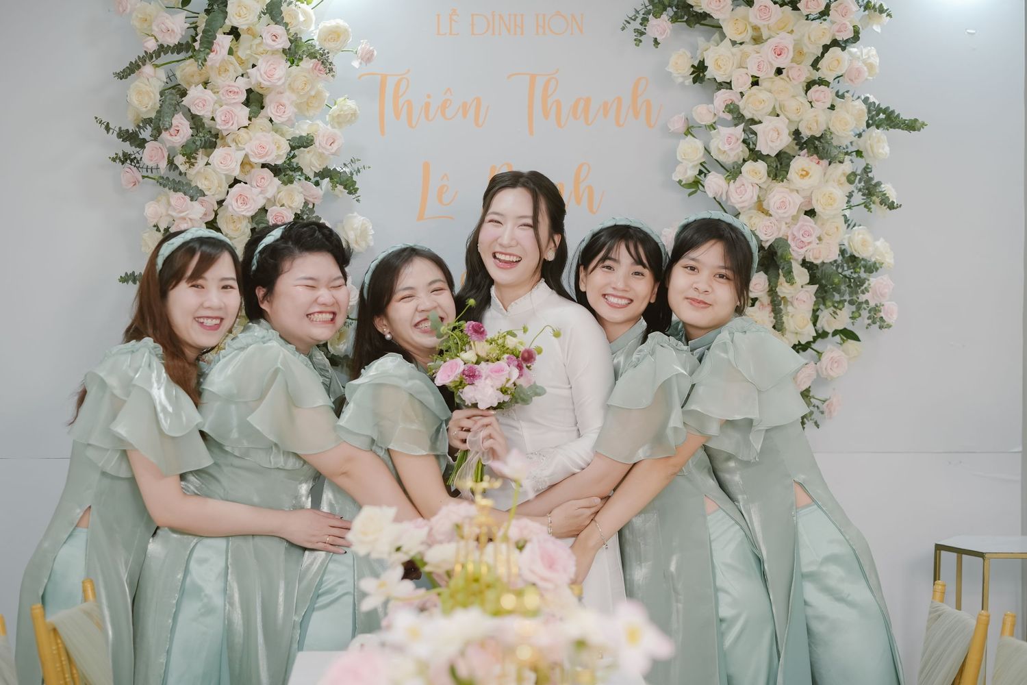 Wedding party in mint green bridesmaids dresses gather around bouquet display with floral backdrop.