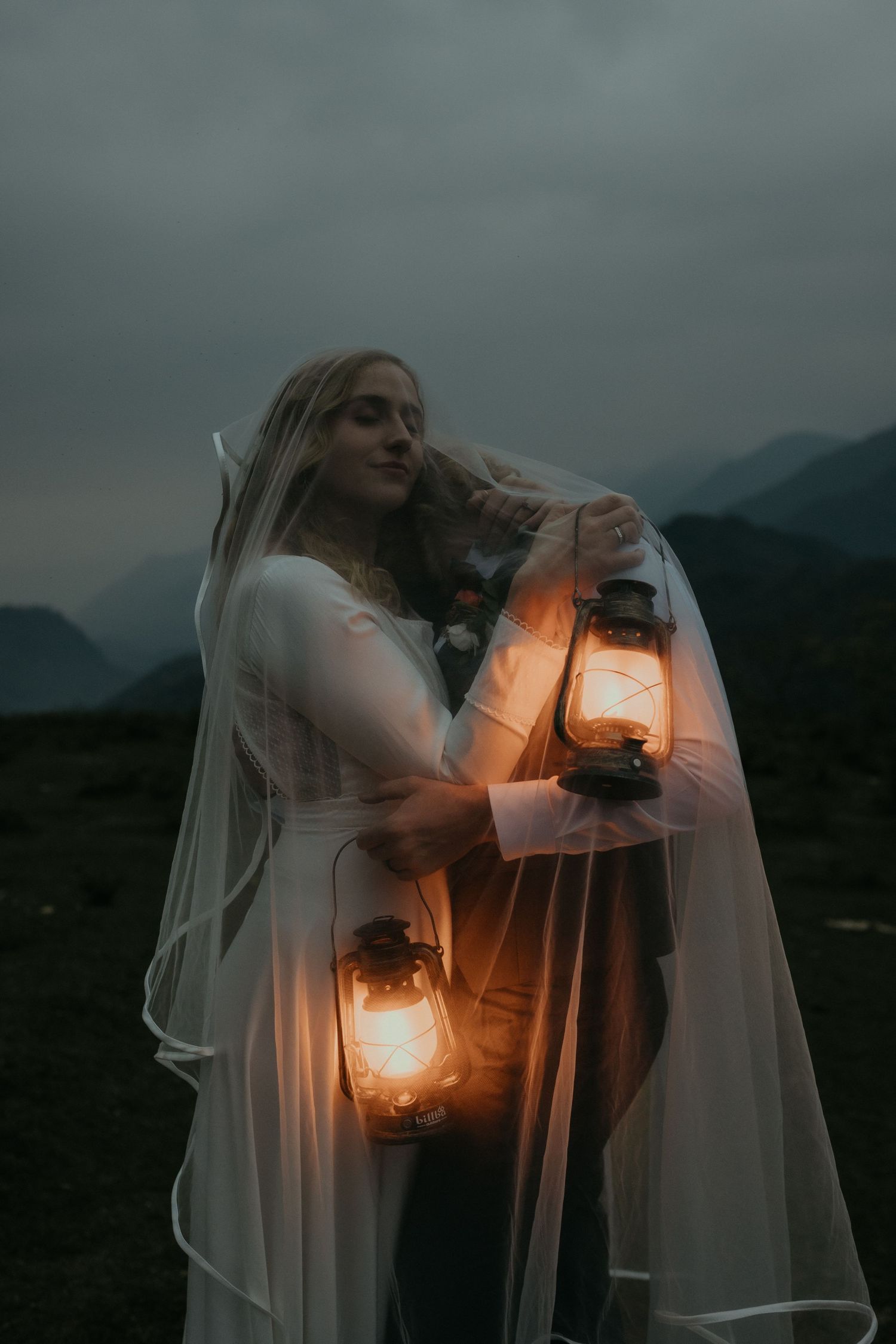A bride holds glowing lanterns in the misty mountain twilight.