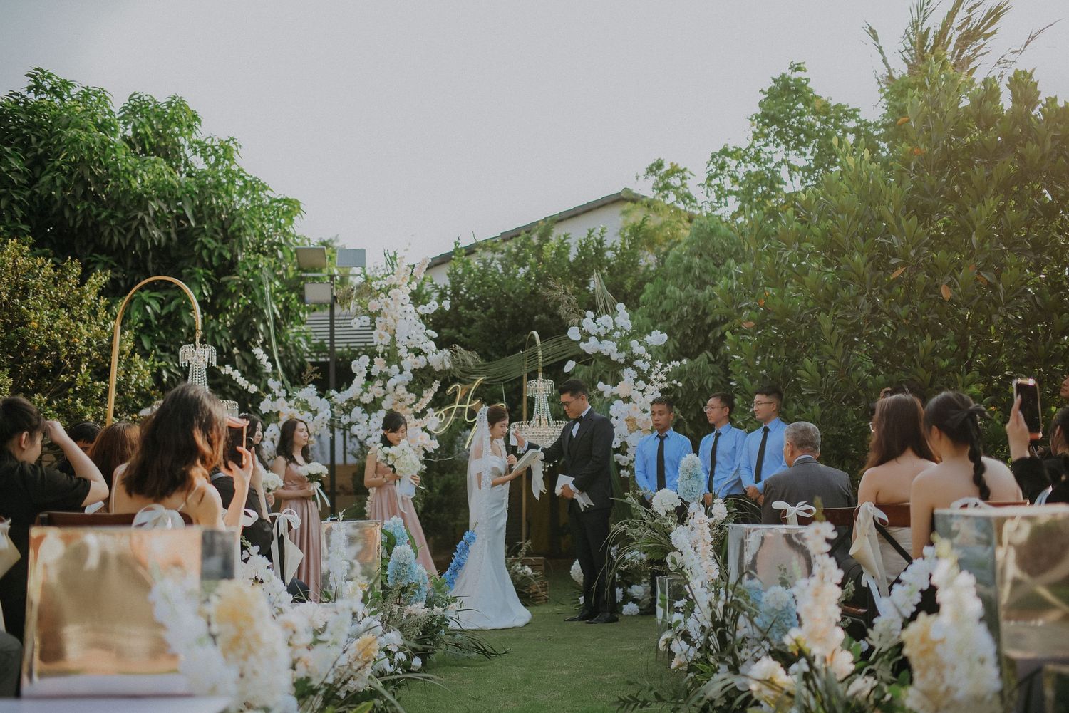 Garden wedding ceremony setup with white floral decorations and arch under trees.